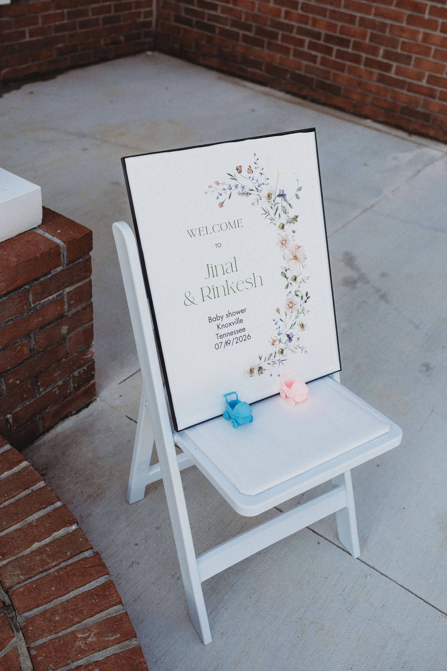 A white folding chair displays a floral welcome sign for Jinal & Rinkesh baby shower, with pink and blue ribbon accents.