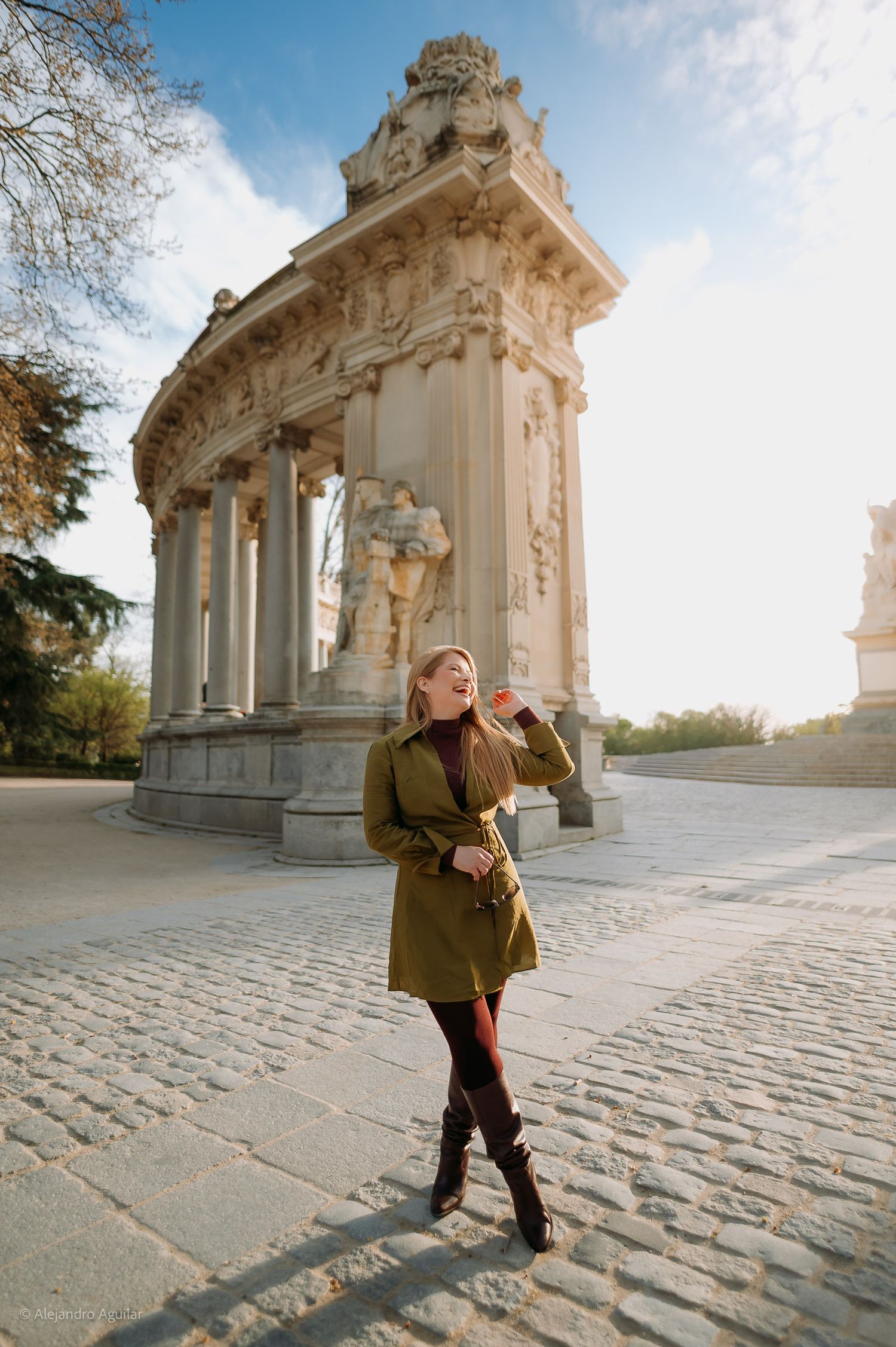 Woman in olive coat laughs joyfully before an ornate baroque monument on a sunny cobblestone plaza.