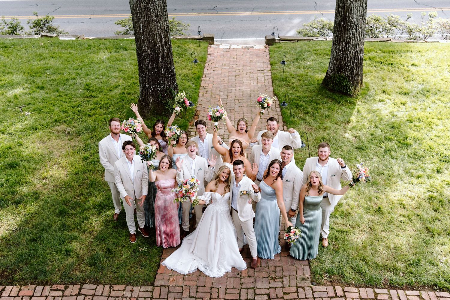 Aerial view of a wedding party celebrating outdoors on a brick path surrounded by lush green grass and trees.