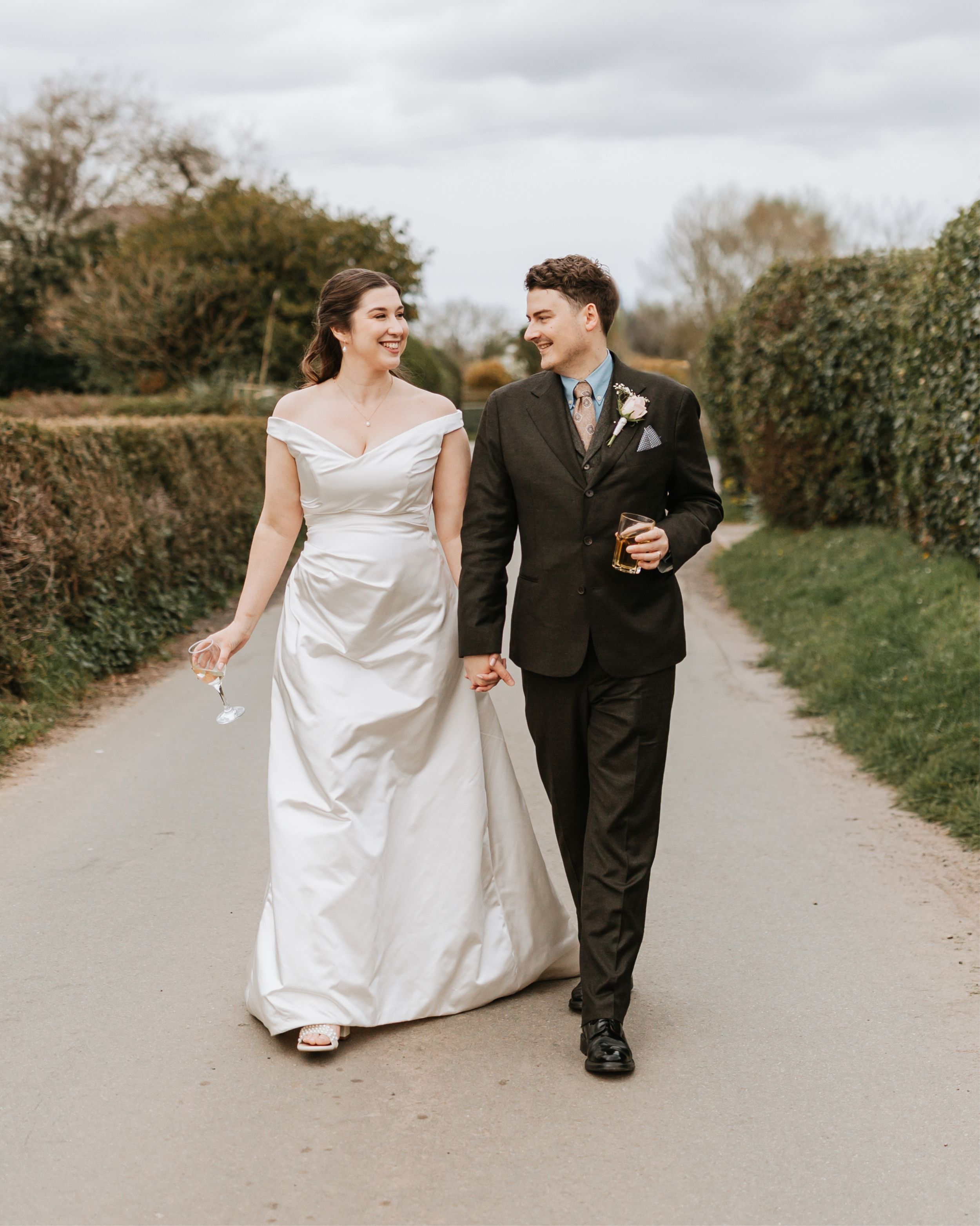 A smiling bride in a white off-shoulder gown walks hand-in-hand with a groom in a dark suit on a country lane.