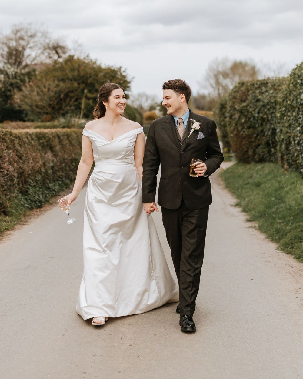 A smiling bride in a white off-shoulder gown walks hand-in-hand with a groom in a dark suit on a country lane.