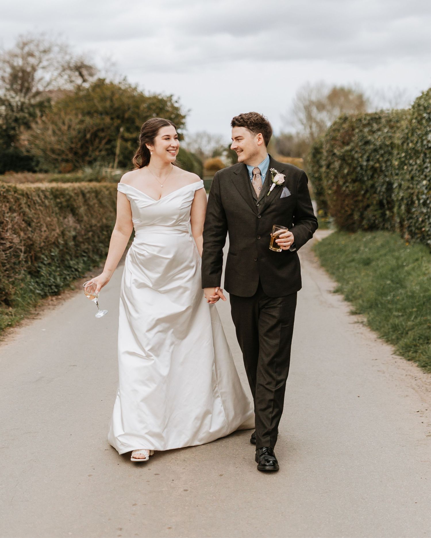 A smiling bride in a white off-shoulder gown walks hand-in-hand with a groom in a dark suit on a country lane.