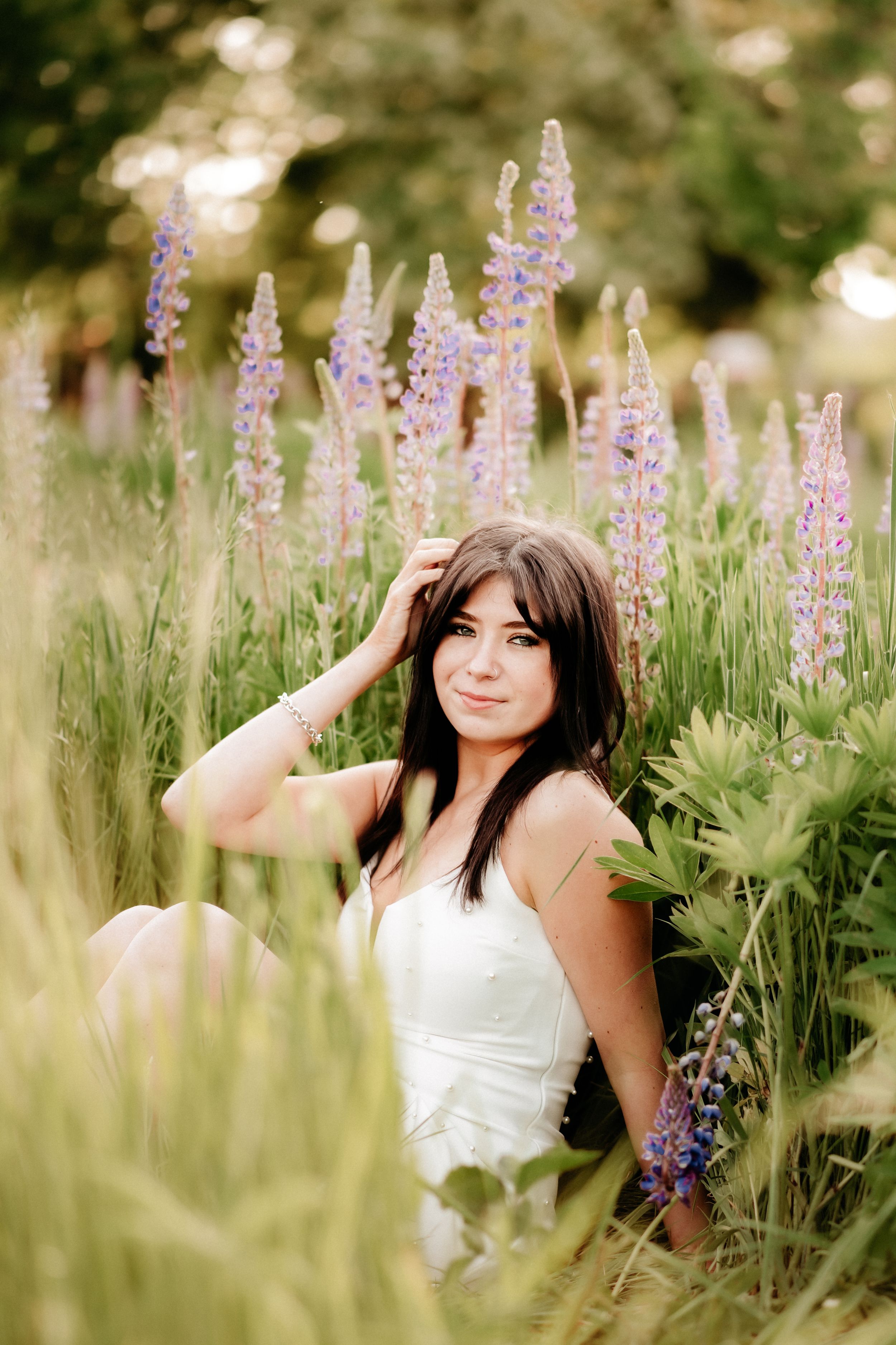 Young woman in white dress sitting among purple lupine flowers in a lush green field, smiling softly at camera for senior photos in Puyallup