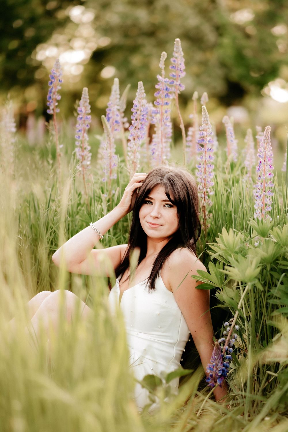 Young woman in white dress sitting among purple lupine flowers in a lush green field, smiling softly at camera for senior photos in Puyallup