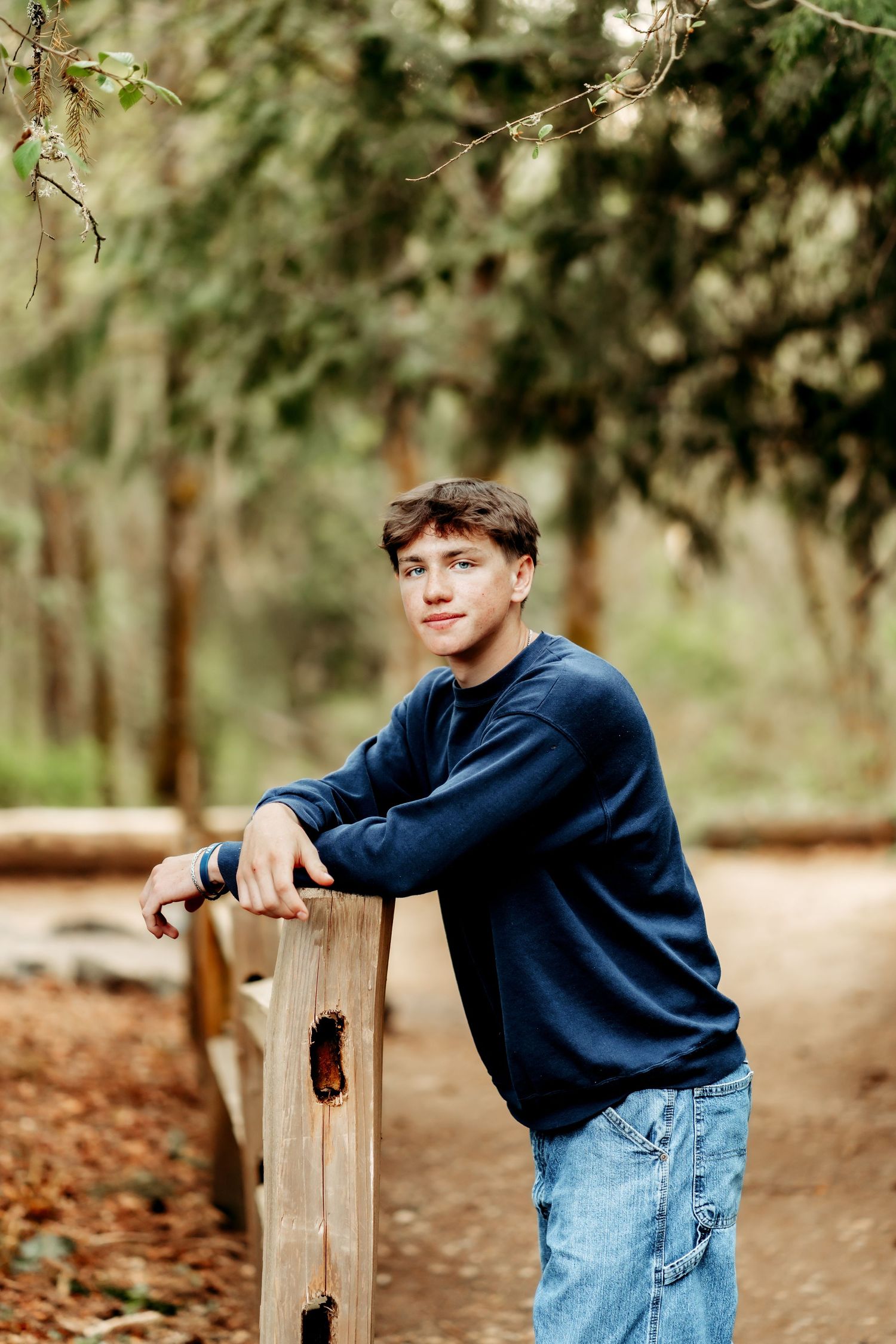 High school senior boy in navy sweater and jeans leans on wooden fence post in lush forest setting during senior photos in Tacoma.