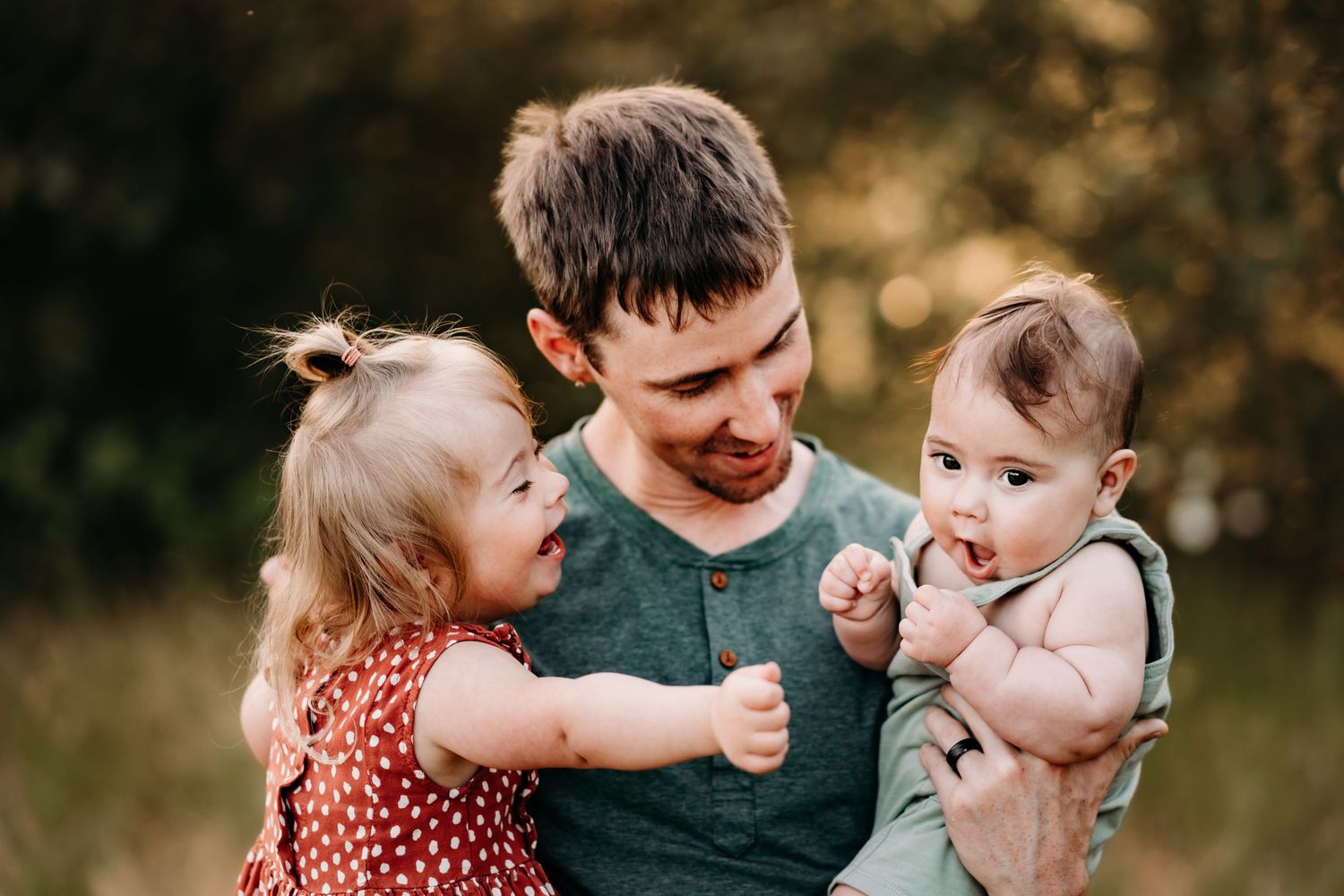 A father holds two young children, a toddler girl in red dress and a baby, outdoors in a sunny field during family photos in Tacoma, WA.