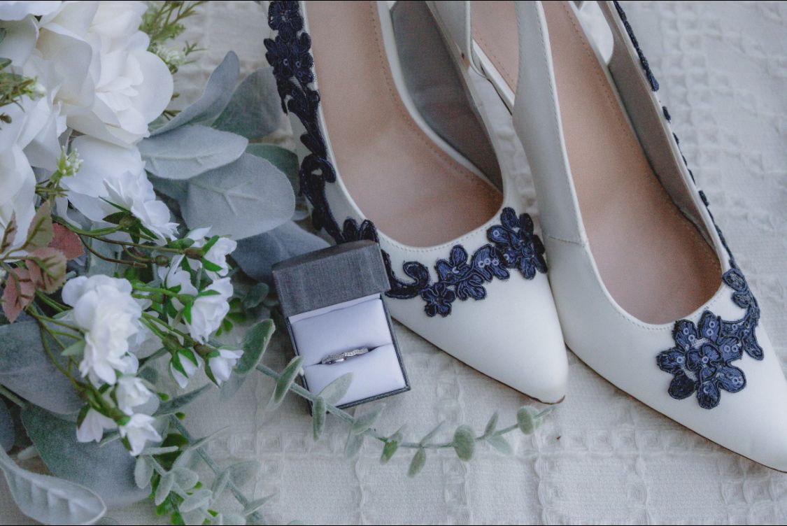 White wedding heels with navy blue lace applique beside a ring box and white floral bouquet.