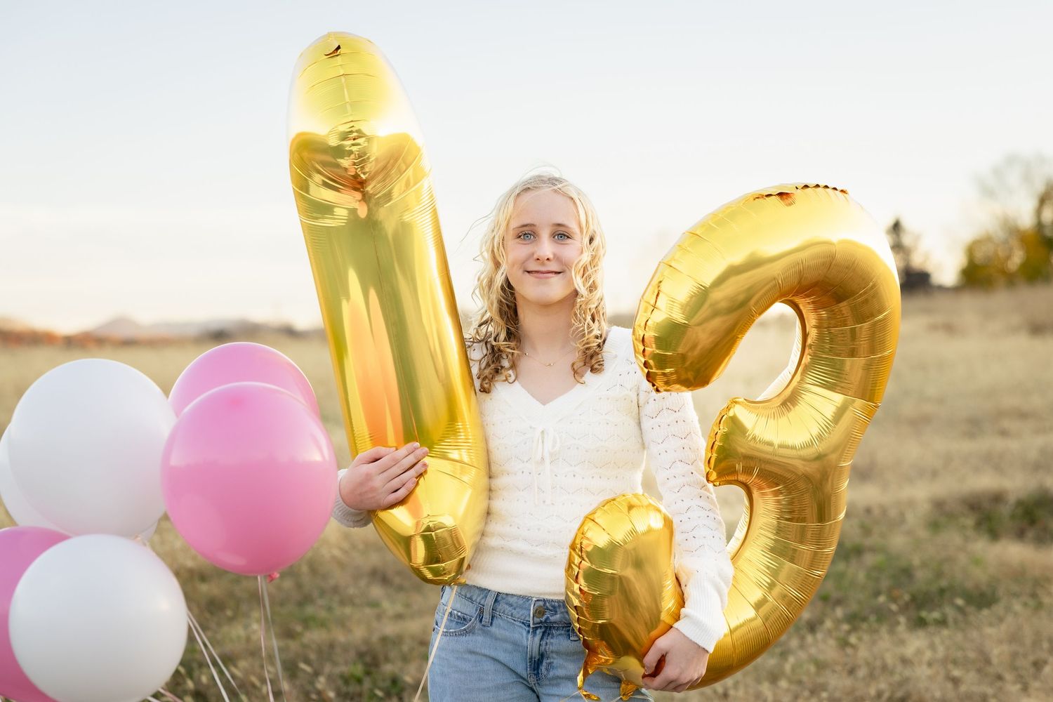 A person holds colorful gold and pink balloons in a golden field during sunset.