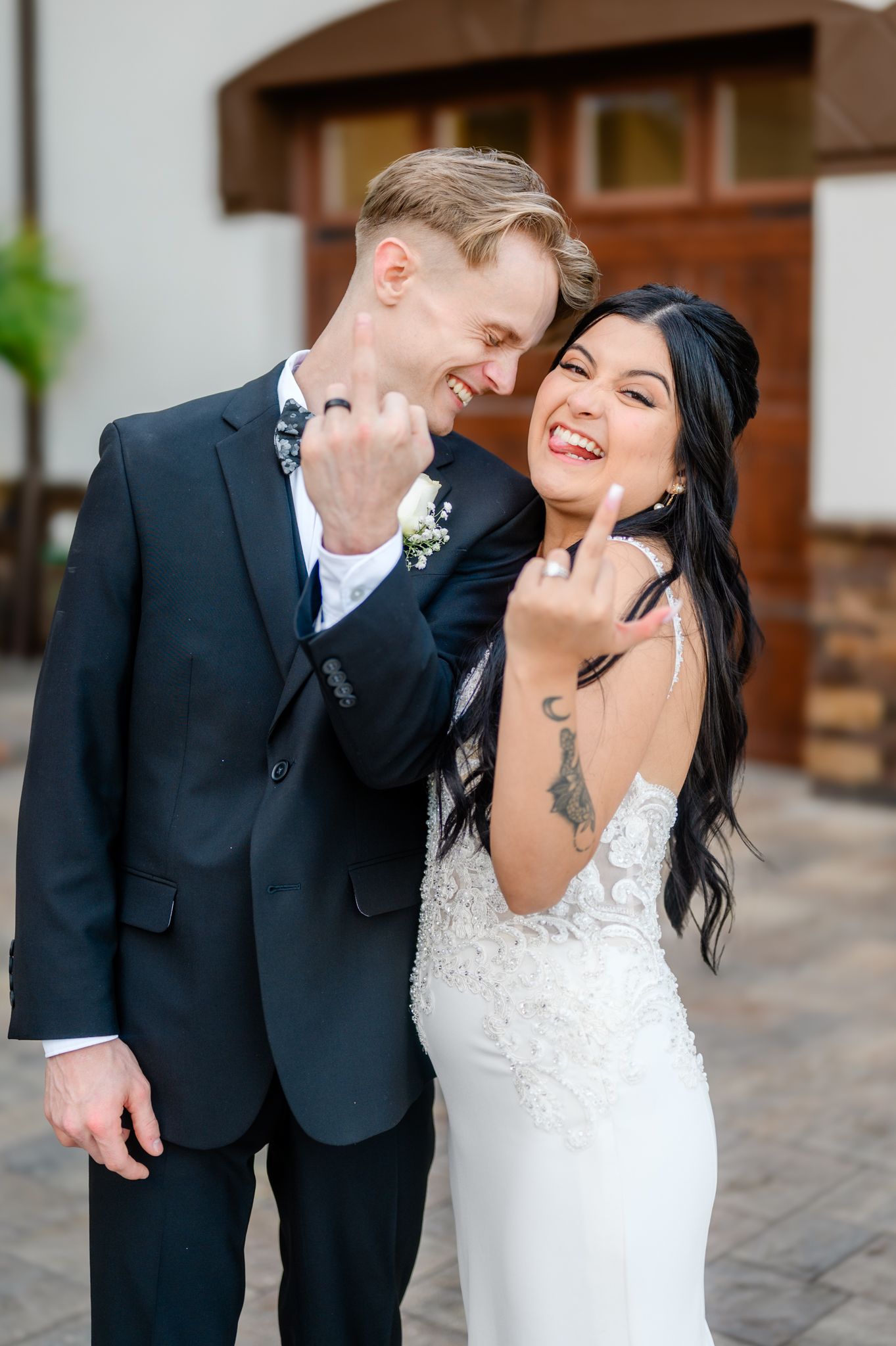 A couple on their wedding day poses in front of a brick archway, with the groom holding a white floral bouquet.