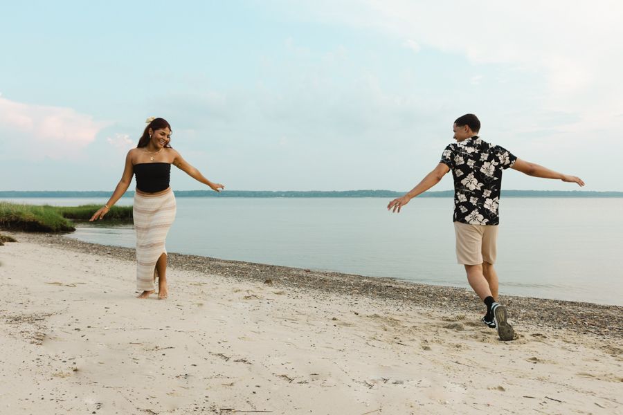 A couple playfully walks along a sandy beach near calm water under a pastel evening sky.