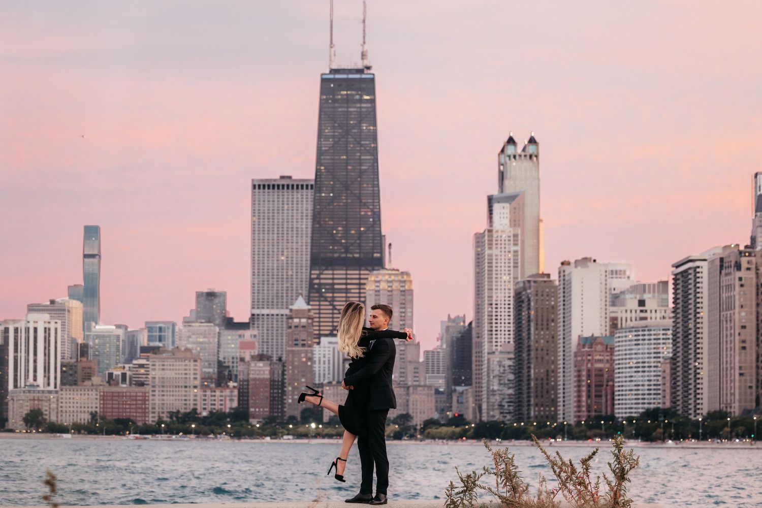 Pink sunset scene of Chicago skyline at sunset with a couple hugging in front of Lake Michigan.