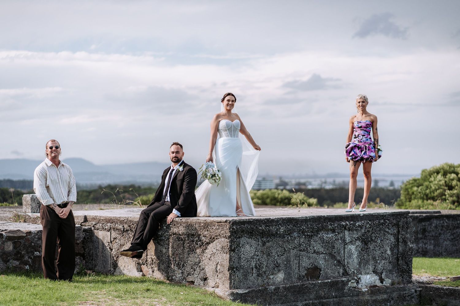 Wedding couple poses on stone wall overlooking scenic coastal landscape during outdoor photo session.