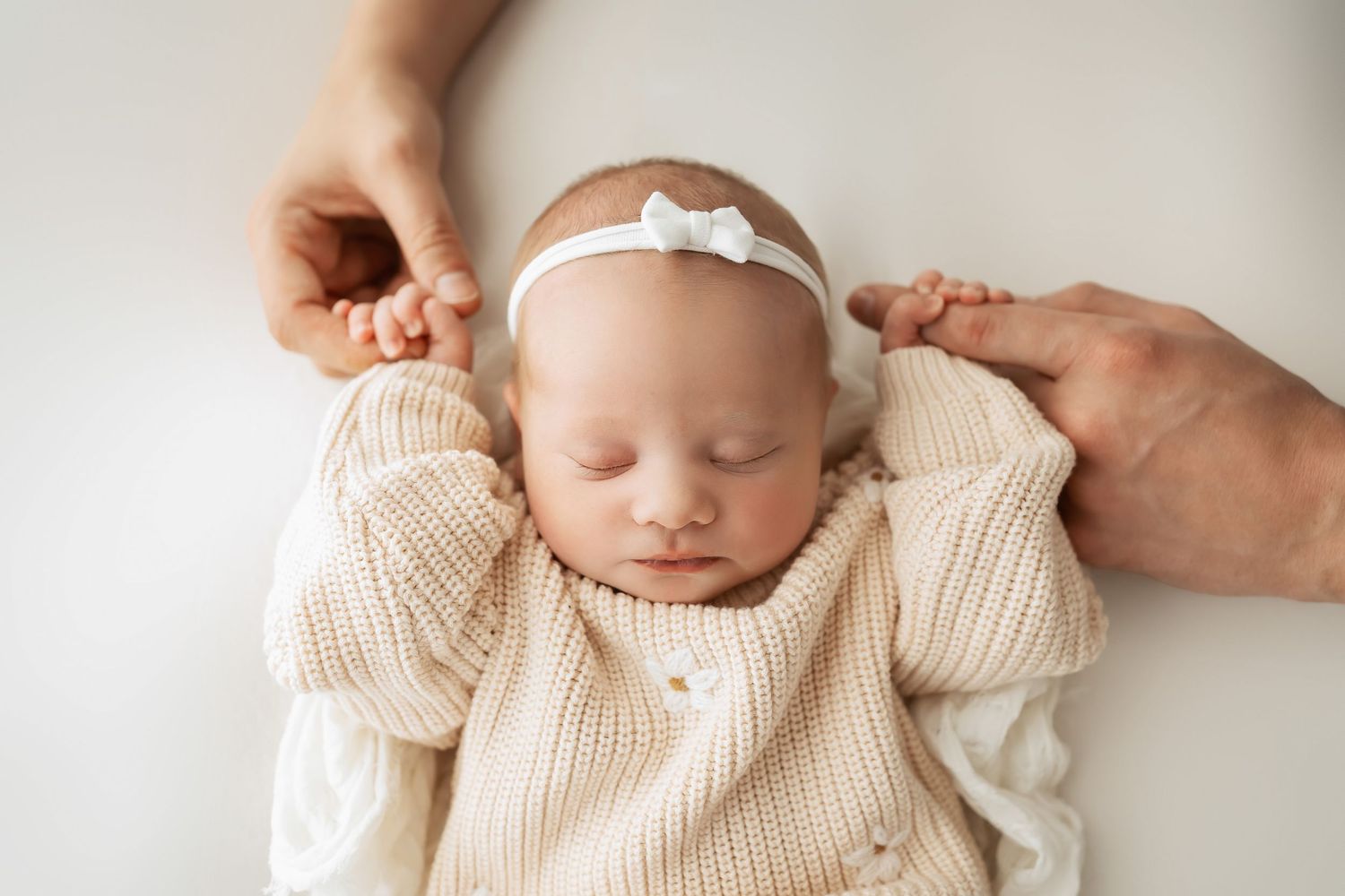 Sleeping newborn wearing cream knit sweater and white bow headband rests peacefully with hands gently held.