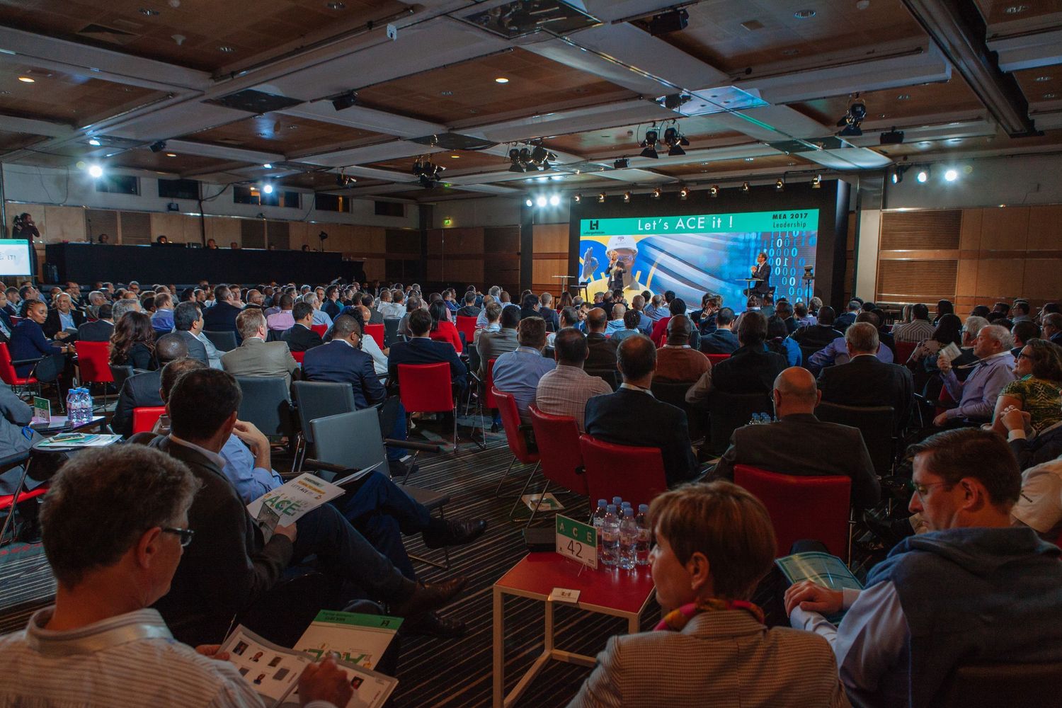 Large audience seated in conference hall watching presentation on screen with red chairs and industrial ceiling.