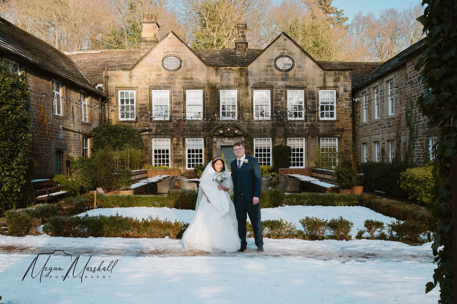 Bride in white gown with veil holding bouquet stands with groom in navy suit on snowy wooden deck surrounded by winter trees.