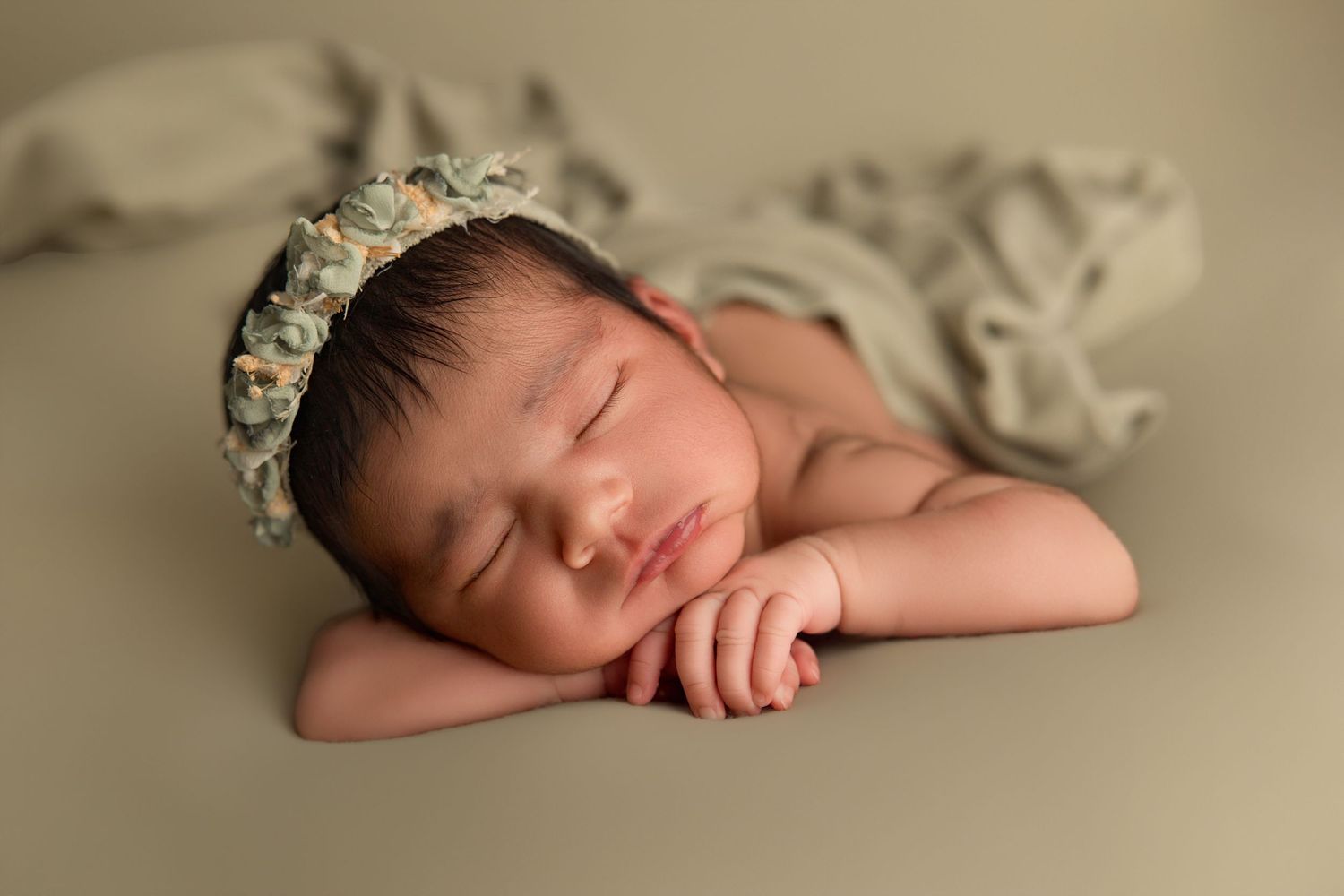 Peaceful newborn baby sleeping with hands tucked under chin, wearing a delicate floral bonnet on a sage green backdrop.