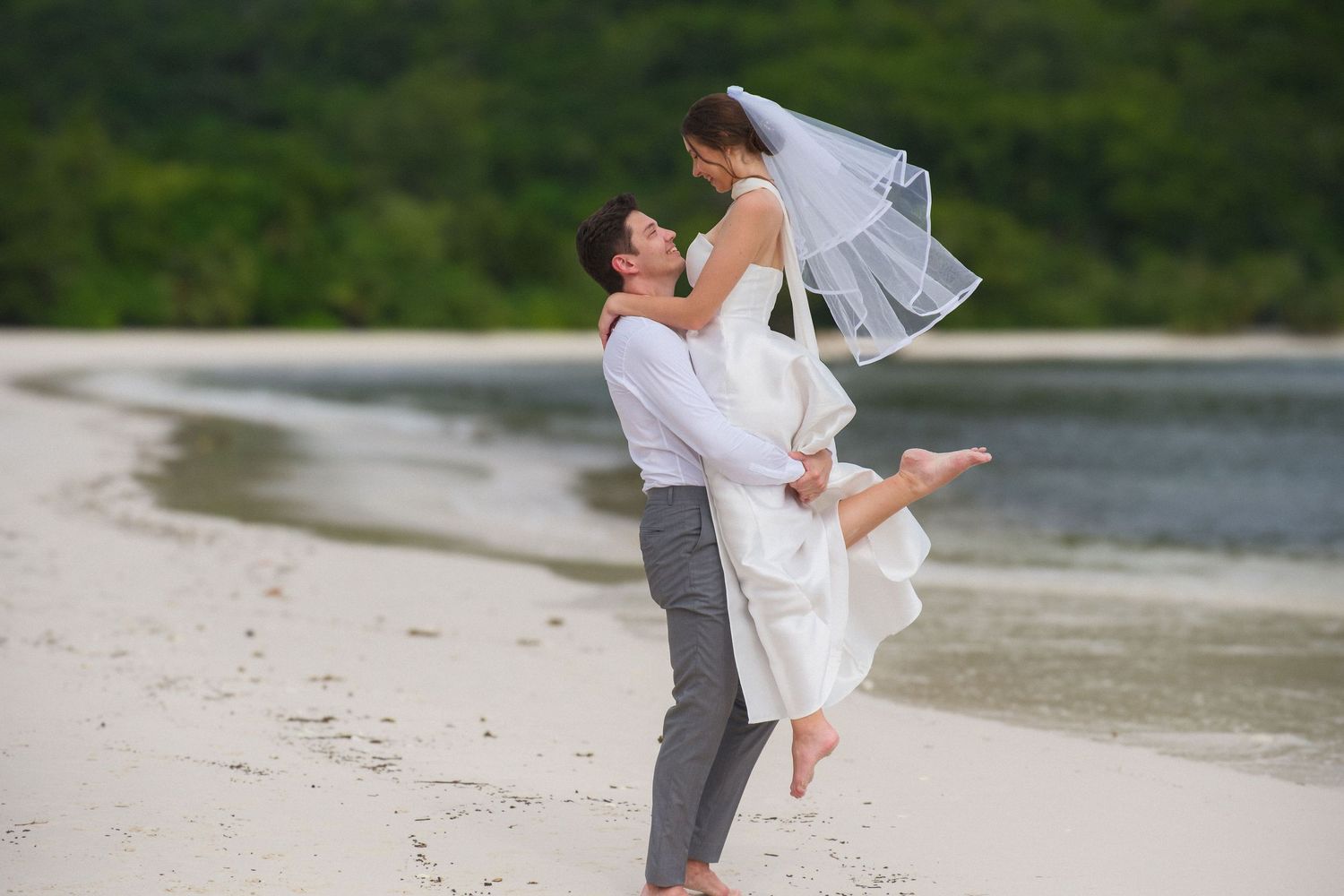 Groom lifting bride in white wedding dress on sandy beach with lush green trees in background.
