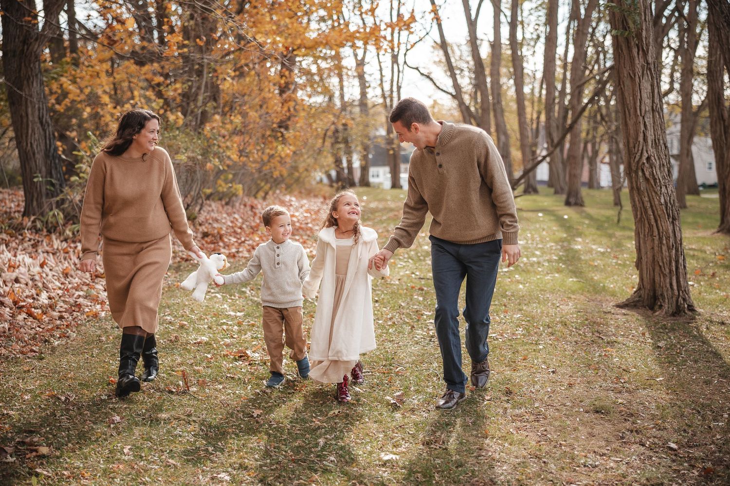 A family walks together through a wooded path with autumn leaves and golden sunlight filtering through the trees.