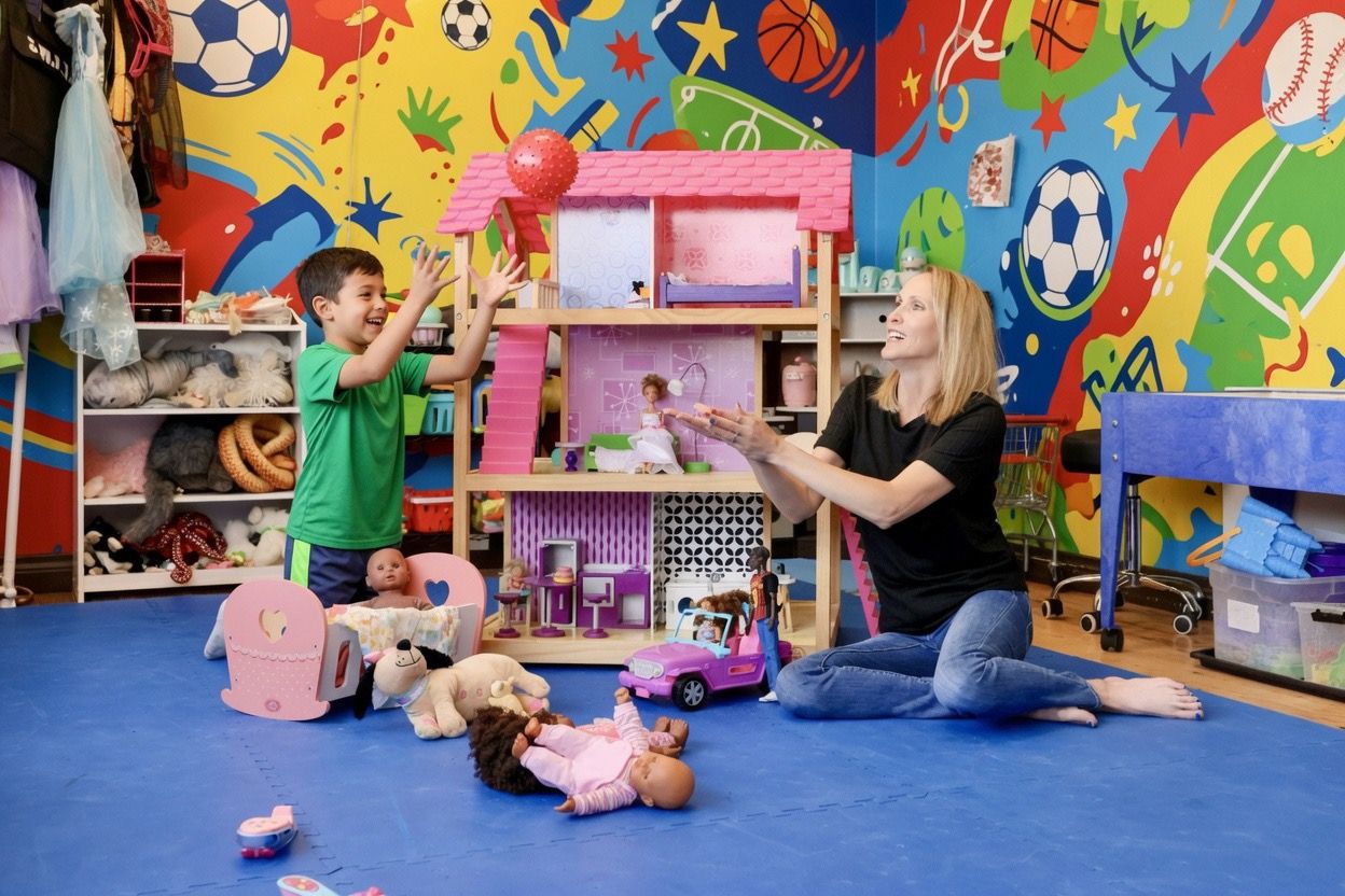 Woman and girl playing with a tall wooden dollhouse surrounded by dolls and toys on a blue floor.