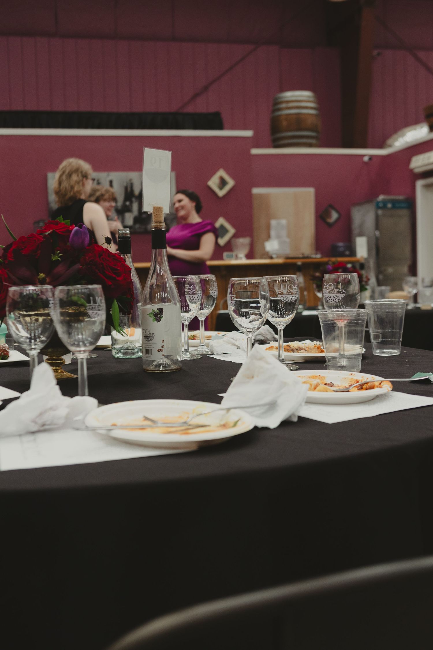 Elegant event table with wine glasses, bottles, and red floral centerpiece in a winery venue with guests in background.