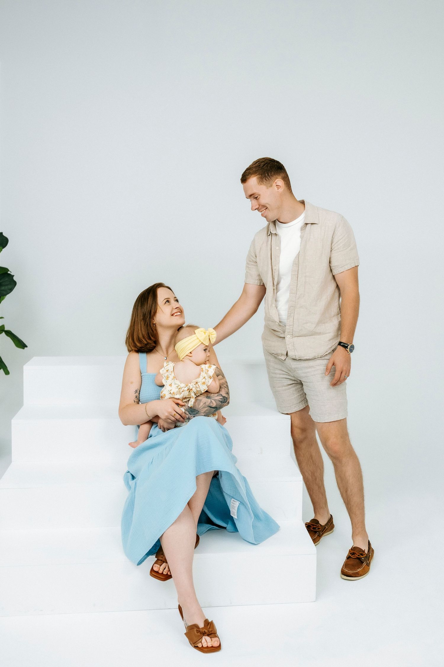A happy family with a mother in a blue dress holding a newborn baby while a smiling father stands beside her on white steps.