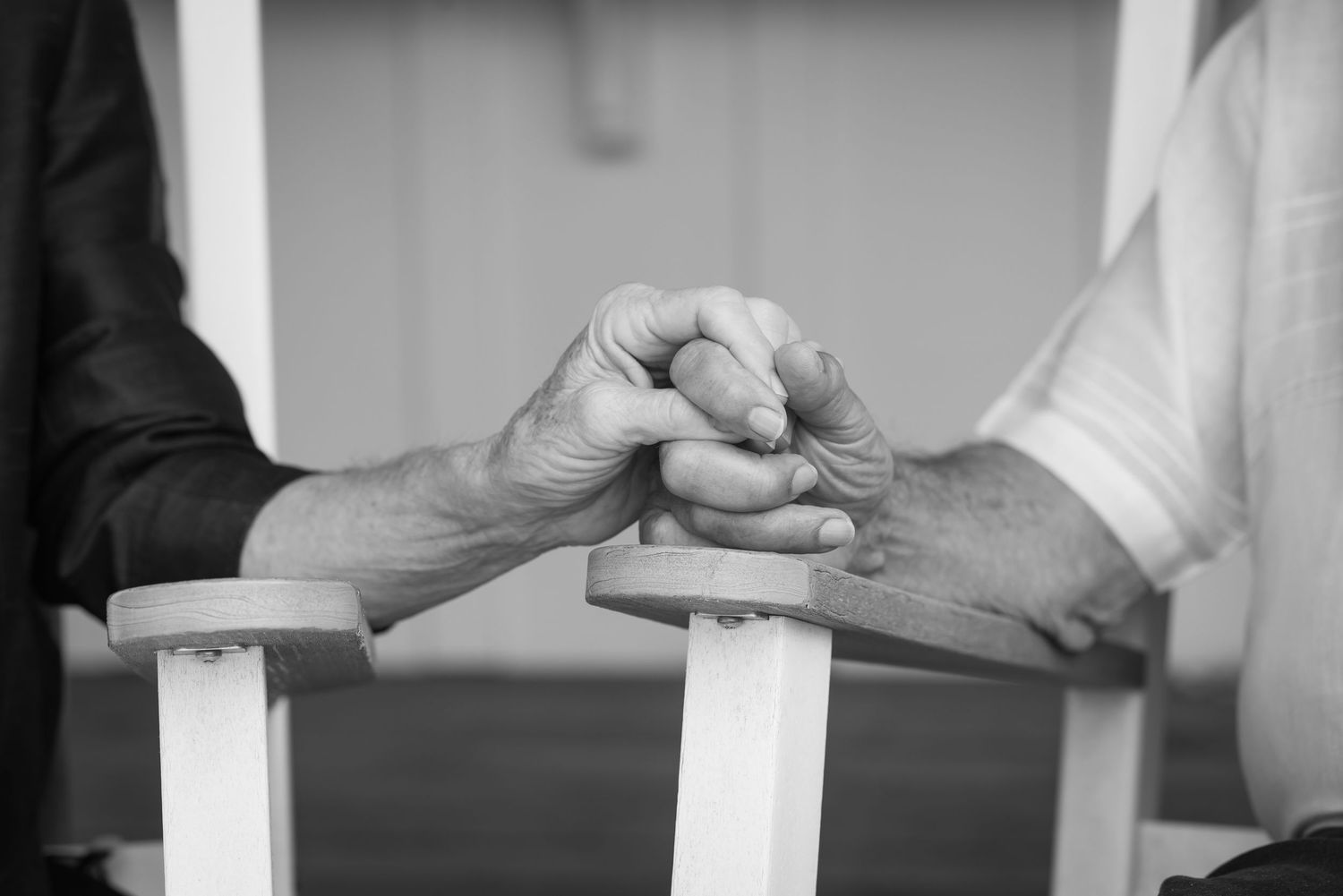 Black and white photo of two elderly people holding hands tenderly while seated in white chairs.