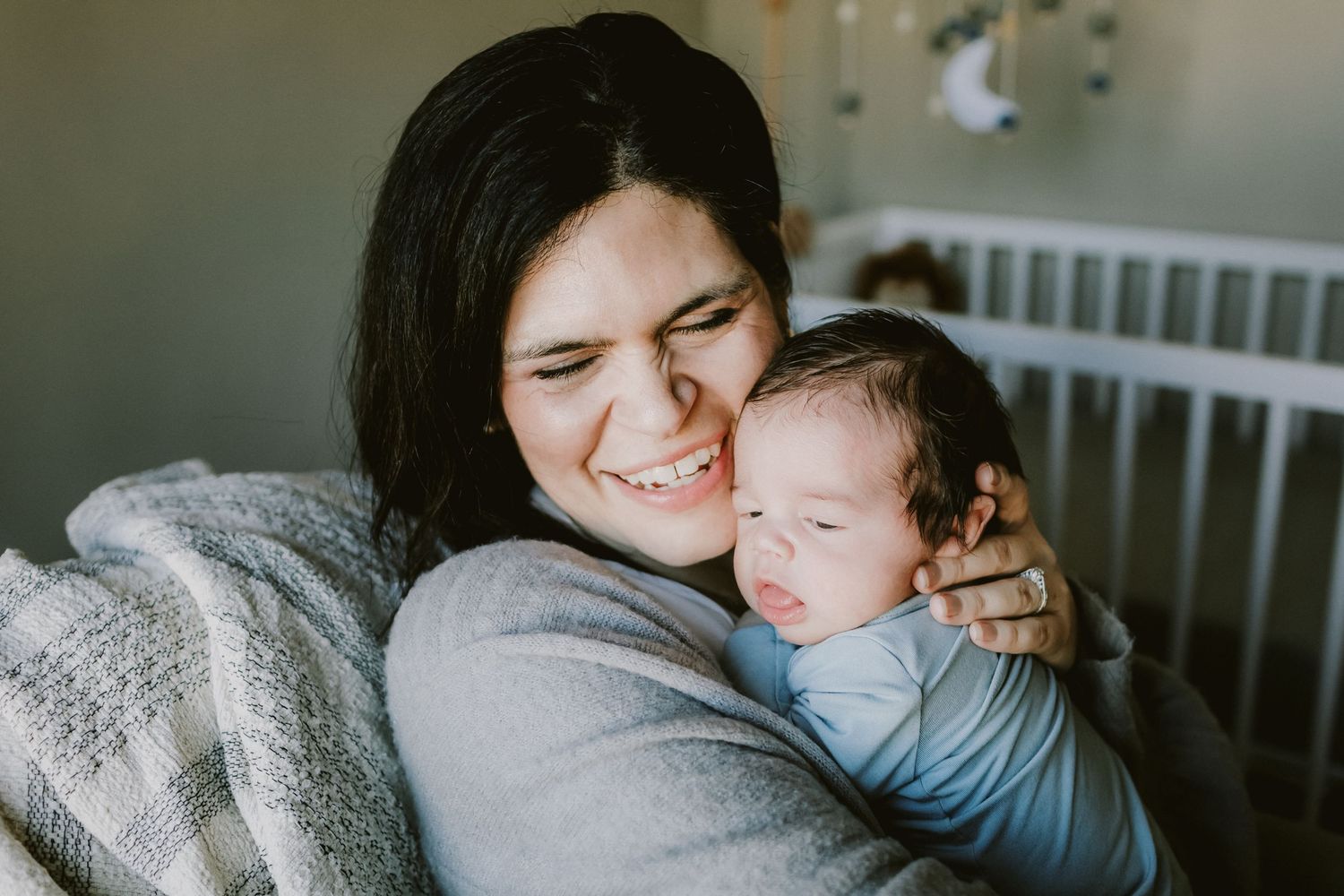 A mother with short dark hair embraces newborn son in the nursery with a very joyful expression for a newborn photoshoot in Haverhill, MA