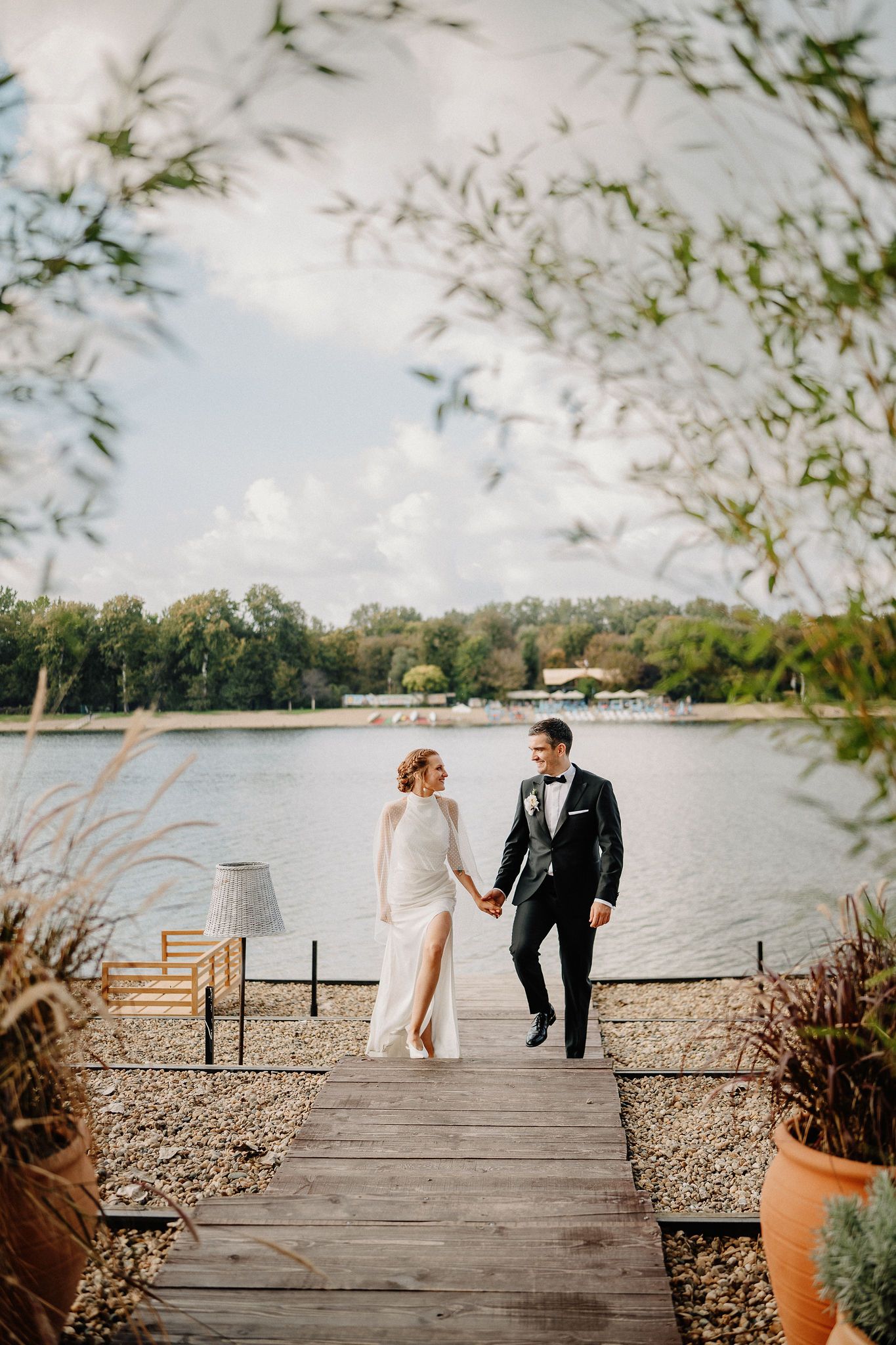 A couple walks hand in hand on a wooden dock overlooking a serene lake surrounded by trees on a cloudy day.