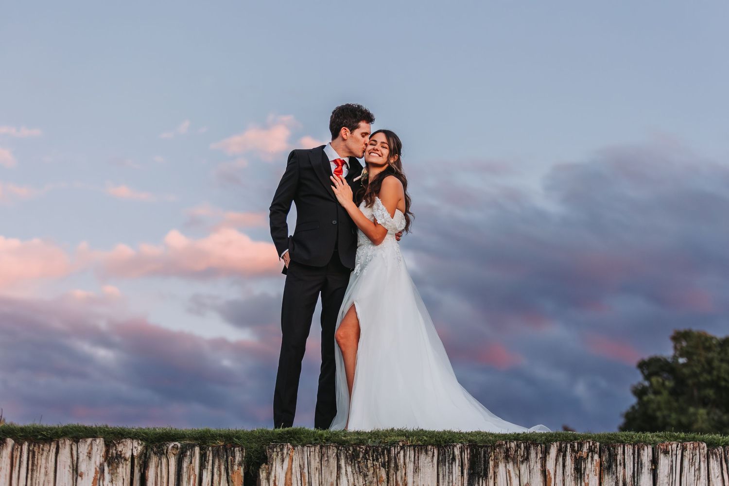 Couple in formal wedding attire embracing on a wooden pier at sunset with dramatic cloudy sky in the background.