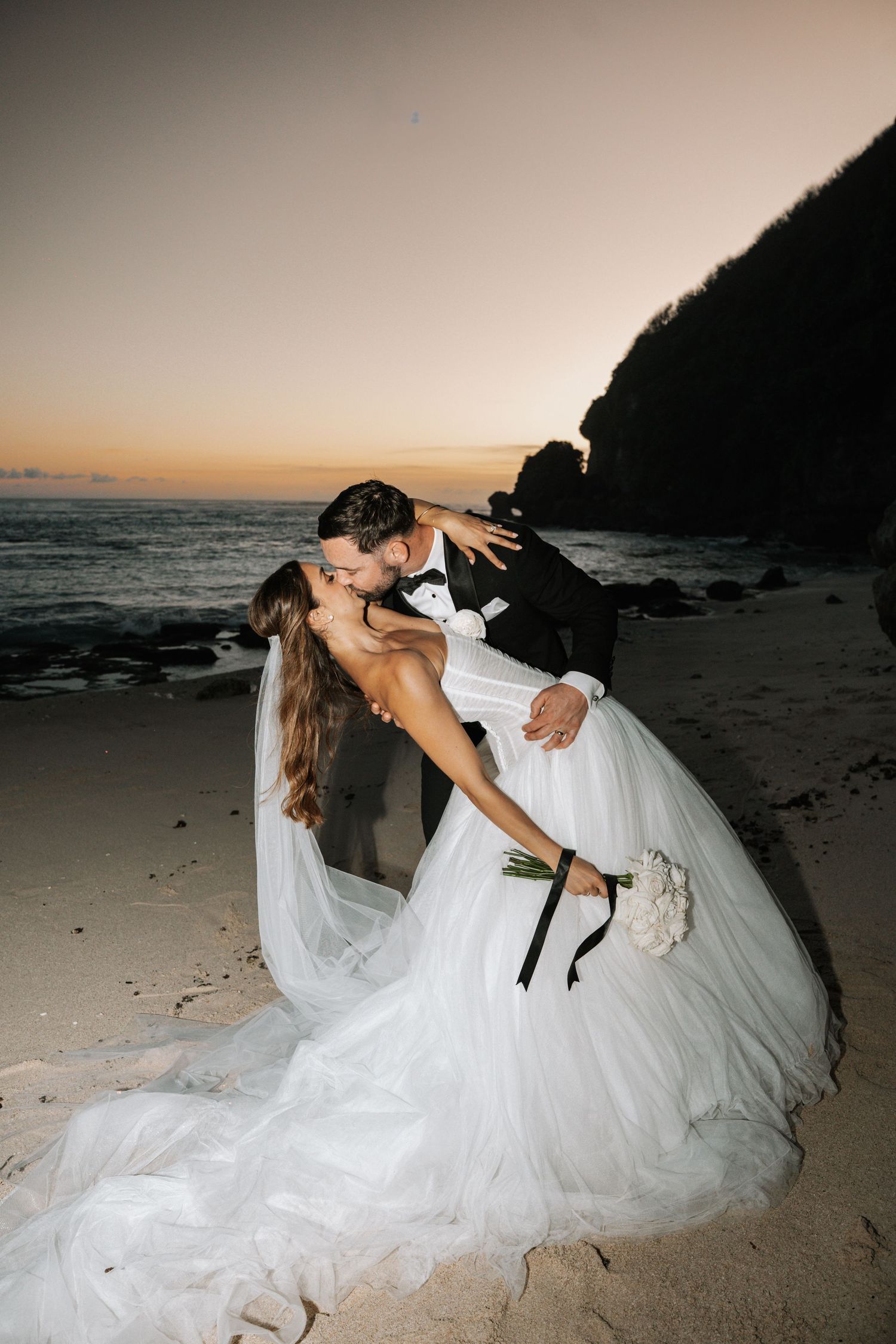 Silhouetted couple sharing a romantic kiss under flowing wedding veil at sunset with dramatic sky in background.