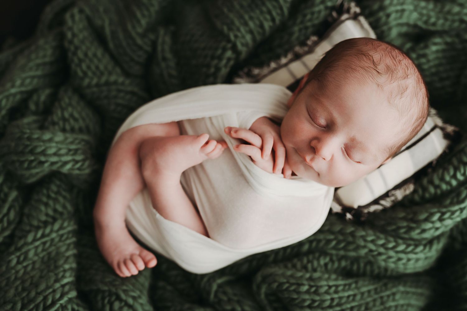 Sleeping newborn baby wrapped in white swaddle blanket lying on textured green knit blanket.