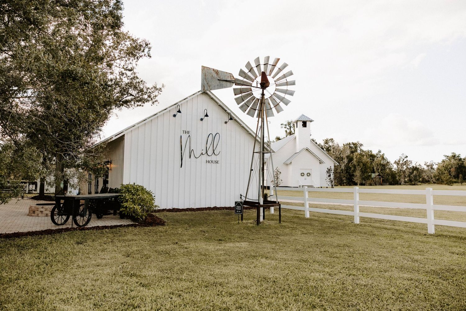 A rustic white barn venue with a vintage windmill sits atop a grassy hill.