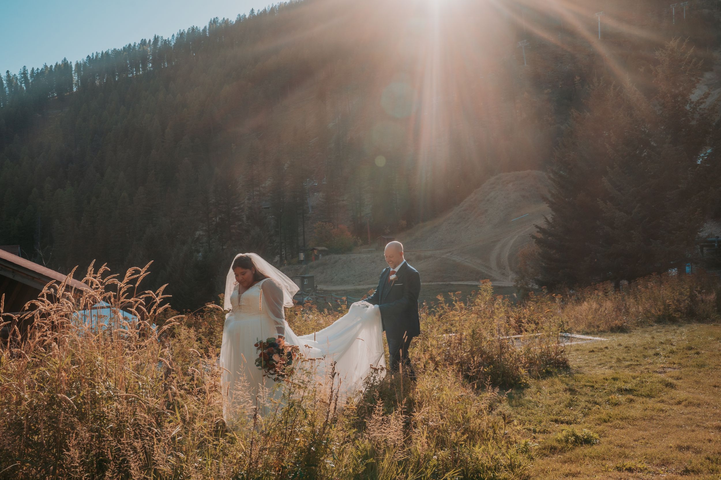 A romantic sunset wedding photoshoot in a golden mountain meadow with sunflare and tall grass.