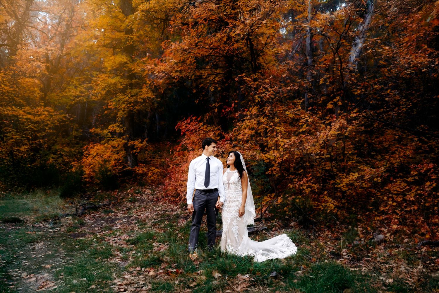 Bride and groom stand together in a vibrant autumn forest with brilliant orange and red fall foliage.