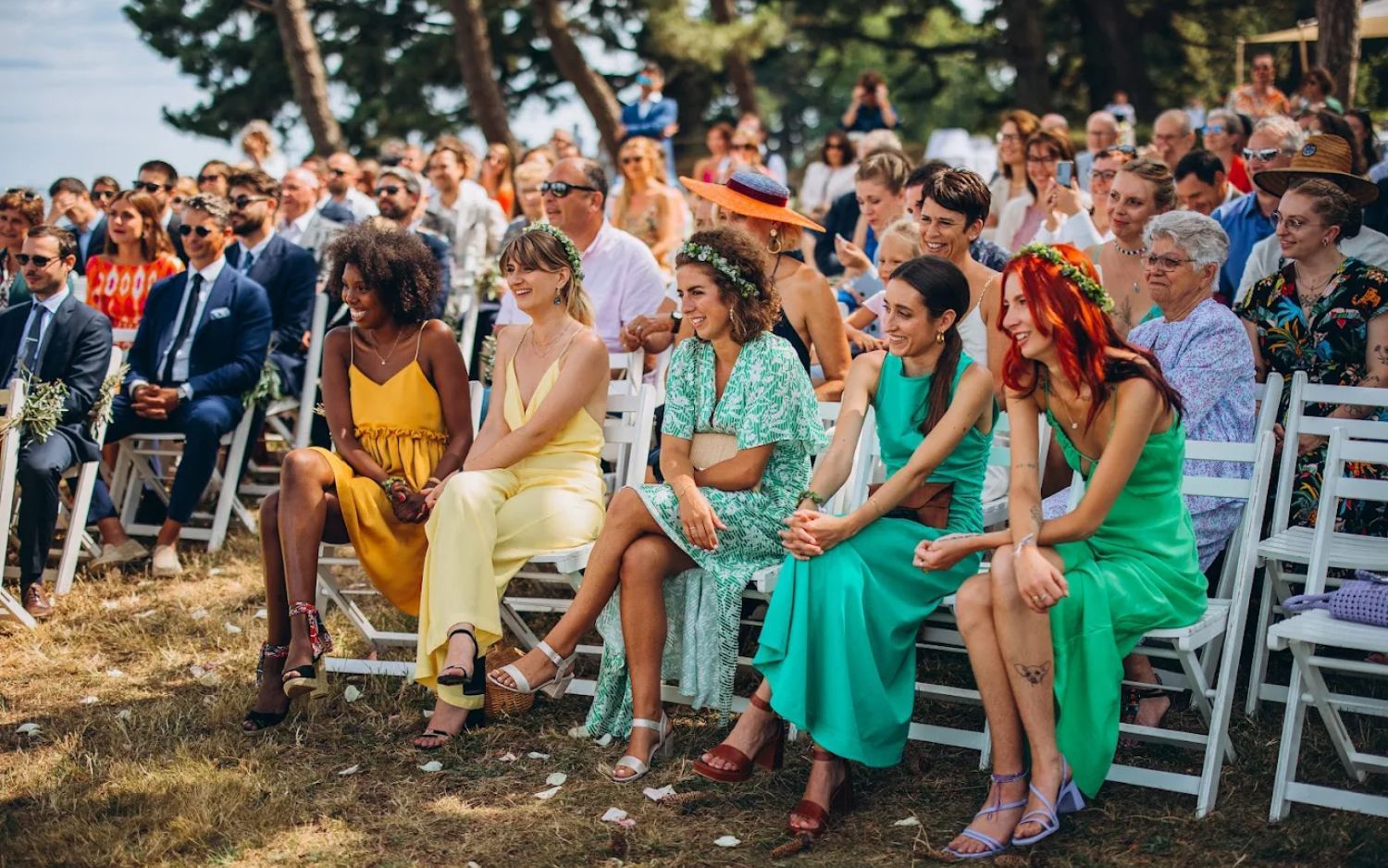 Wedding guests sitting in white chairs during an outdoor summer ceremony with natural lighting and greenery.