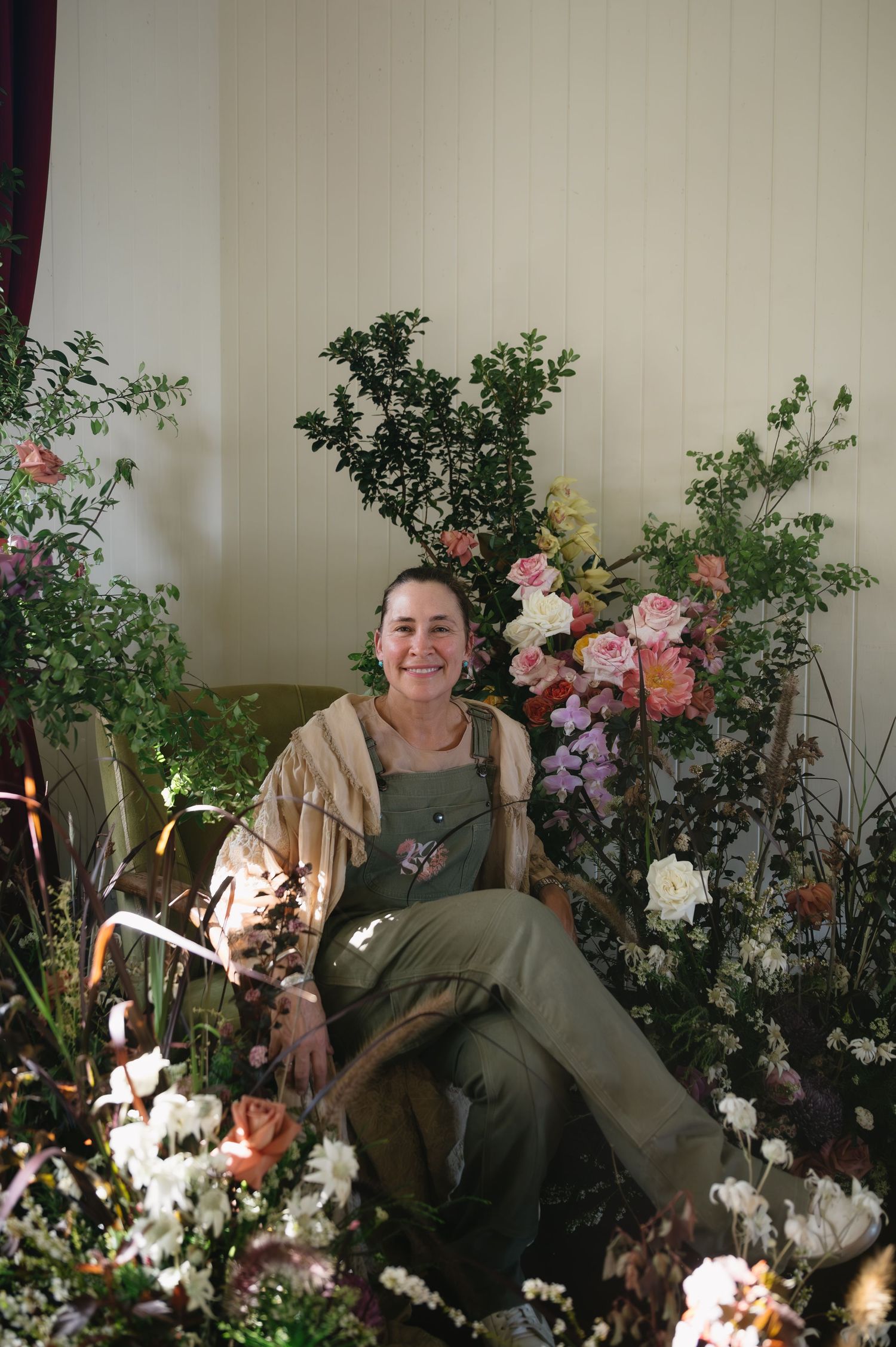 A person lounges in a garden setting surrounded by pink and white flowers and lush greenery against a light wall.