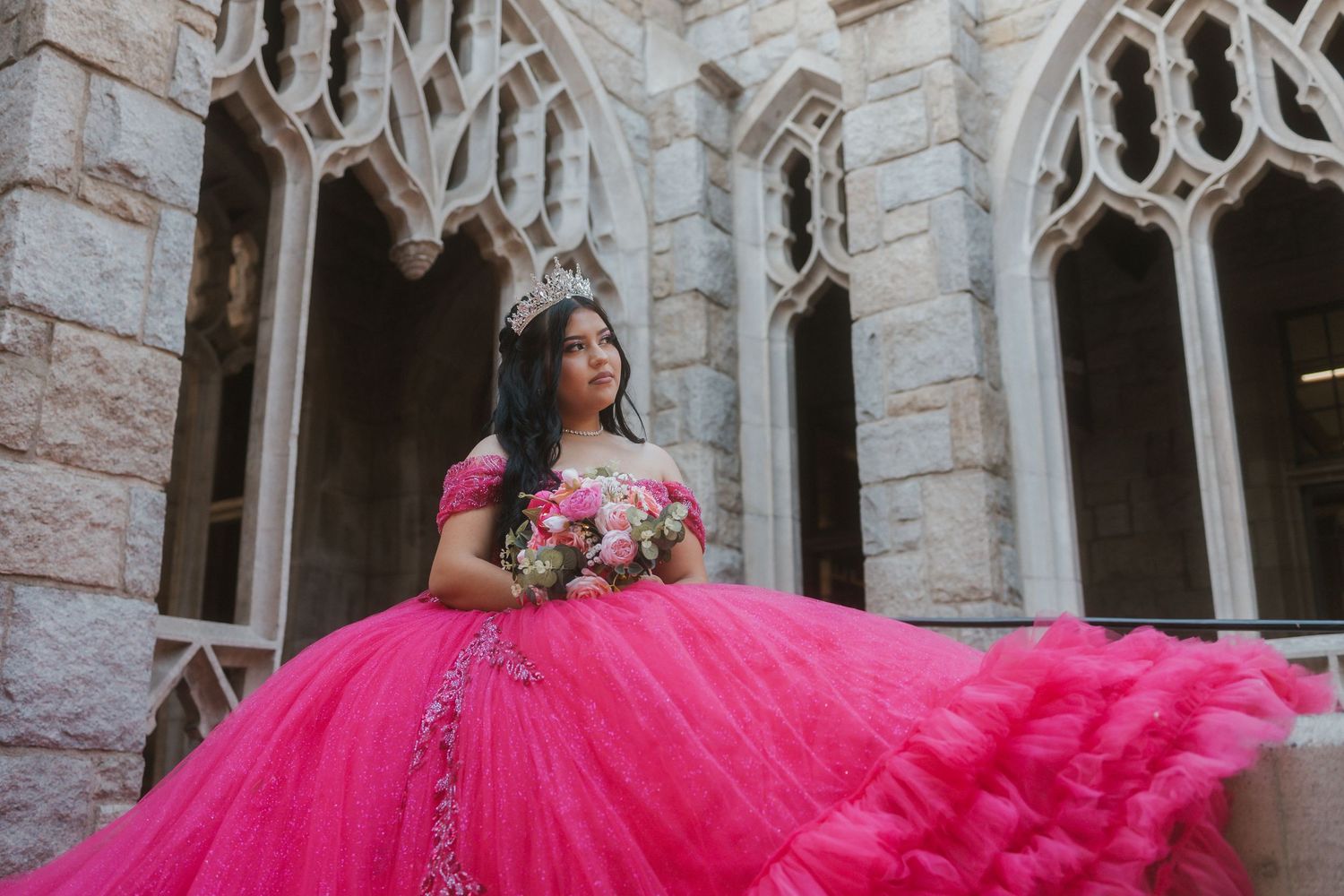 Someone in a vibrant pink quinceanera dress poses elegantly in an ornate stone archway.