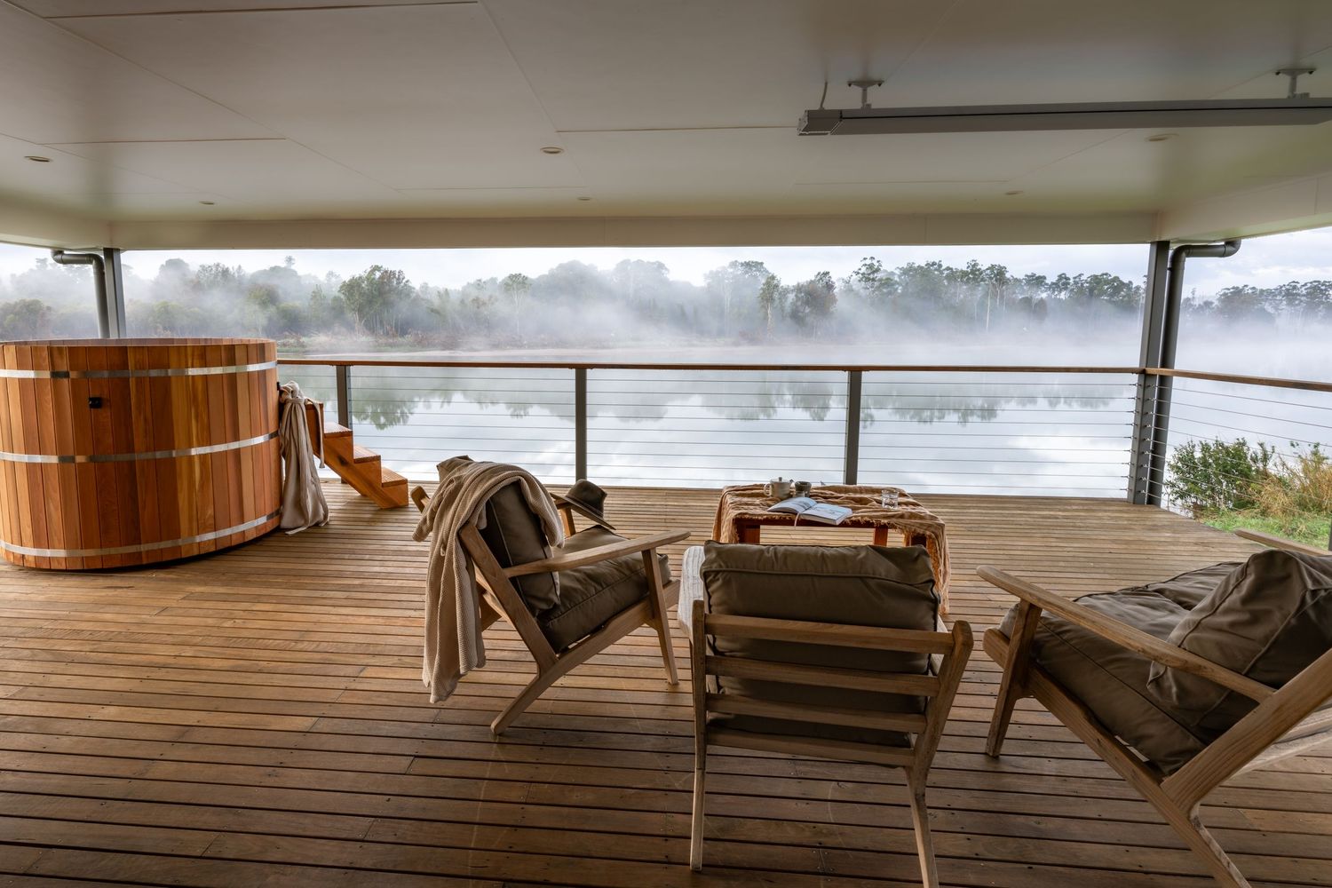 Wooden lounge chairs overlook a misty lake from a covered deck at dawn.