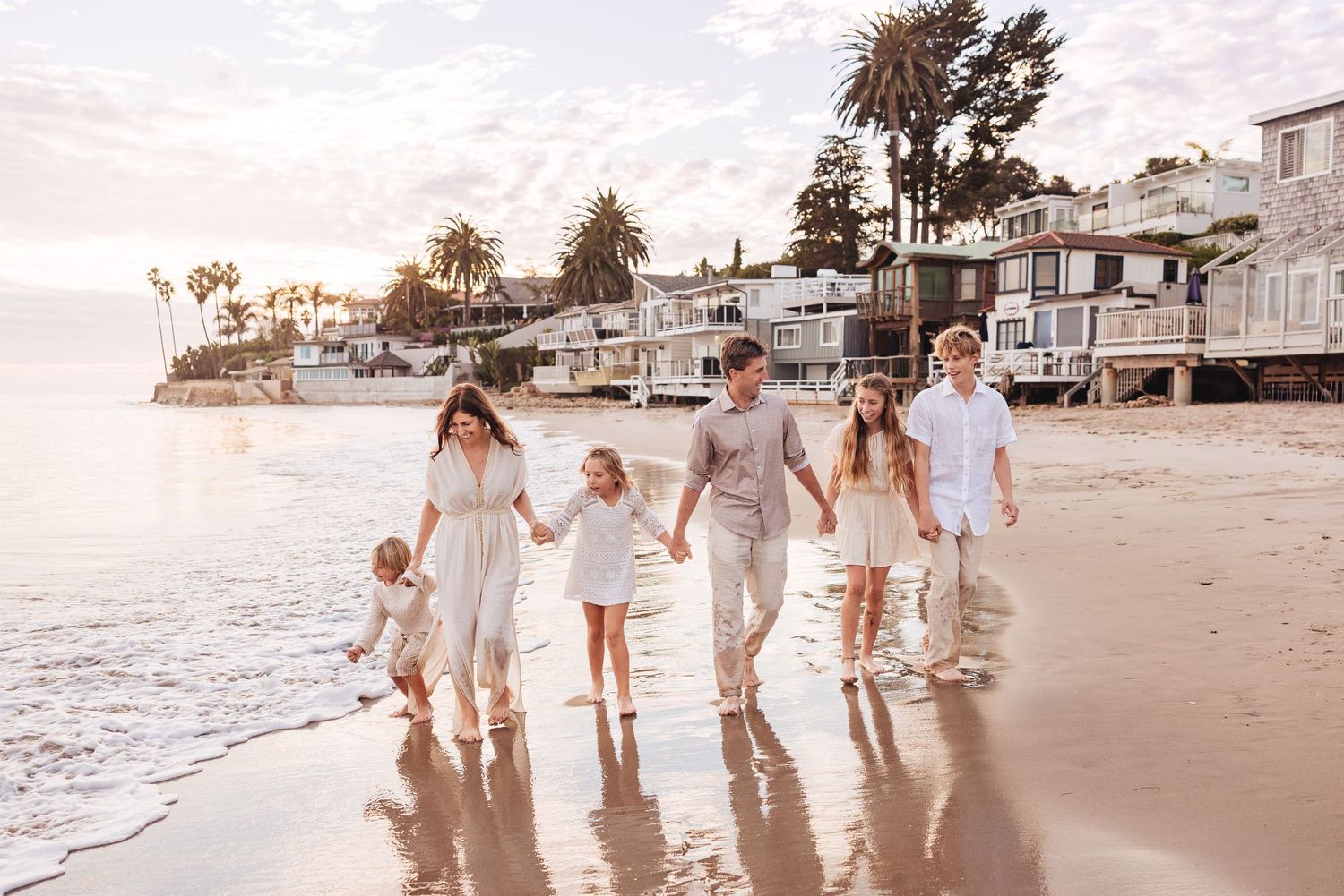 A group in light-colored clothing walks together along a sandy beach at sunset with beachfront houses in the background.