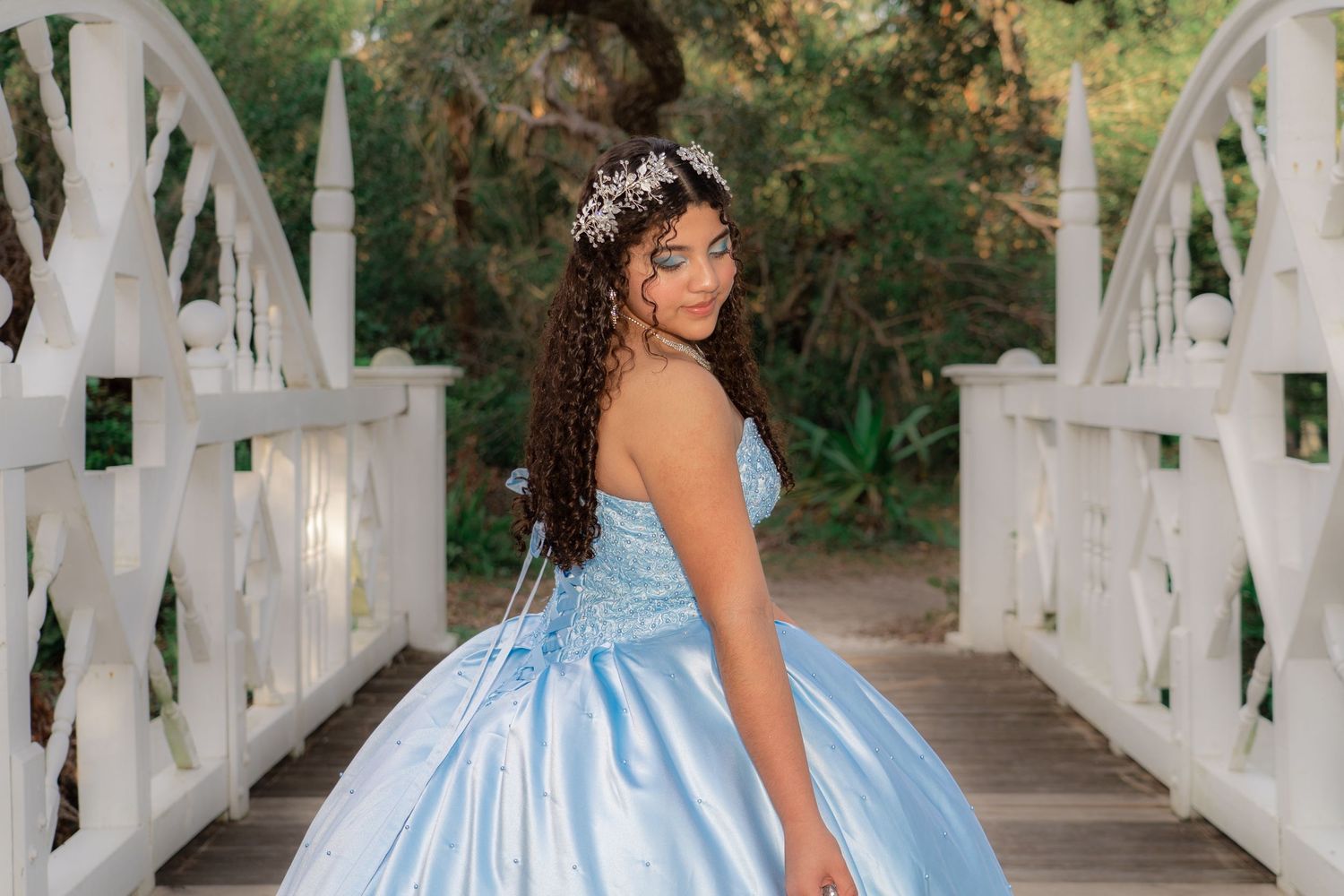 Teen girl in light blue quinceañera gown with floral crown posing on white garden bridge.