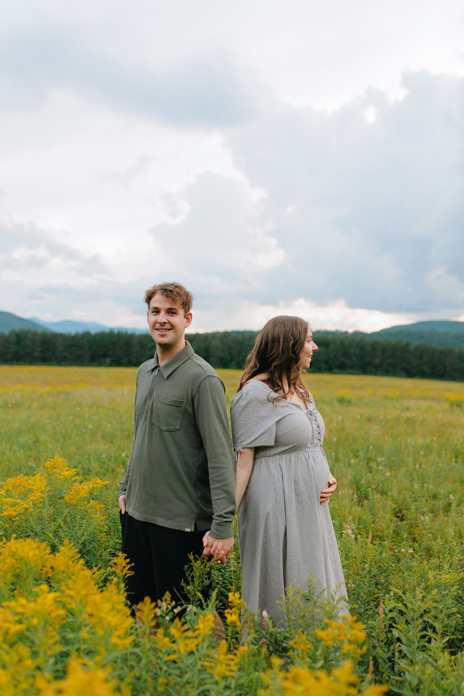 A couple stands back to back in a scenic field of golden wildflowers with mountains visible in the cloudy background.