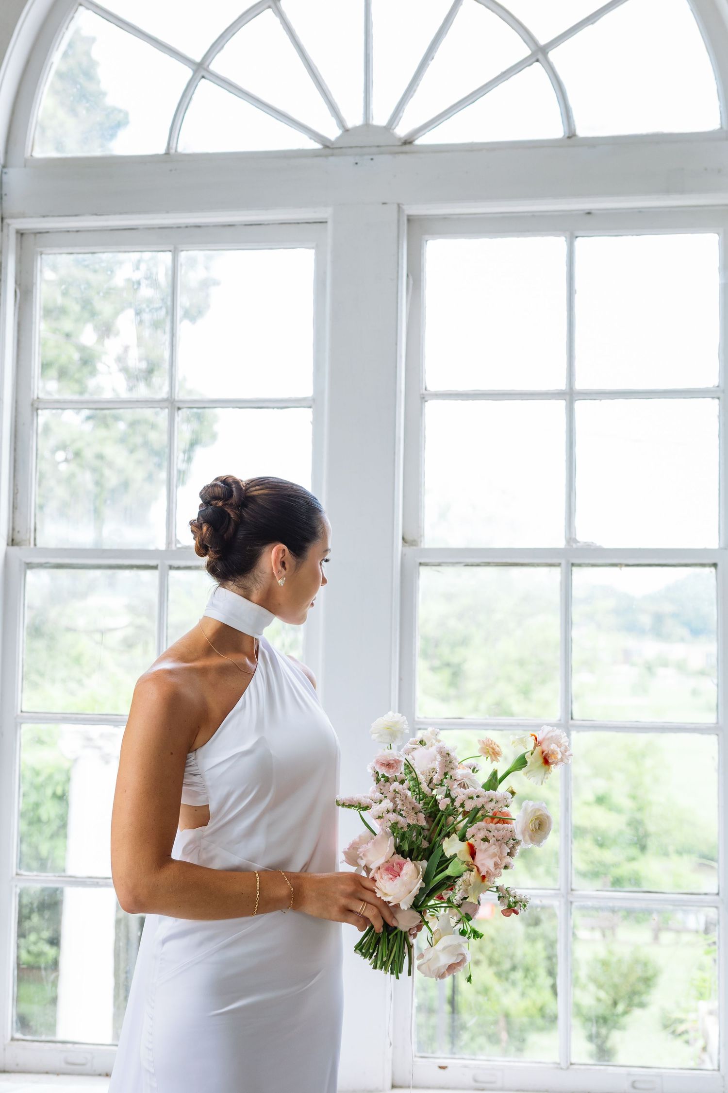 Bride holding pink and white floral bouquet stands in bright sunroom with multiple windows and natural light.