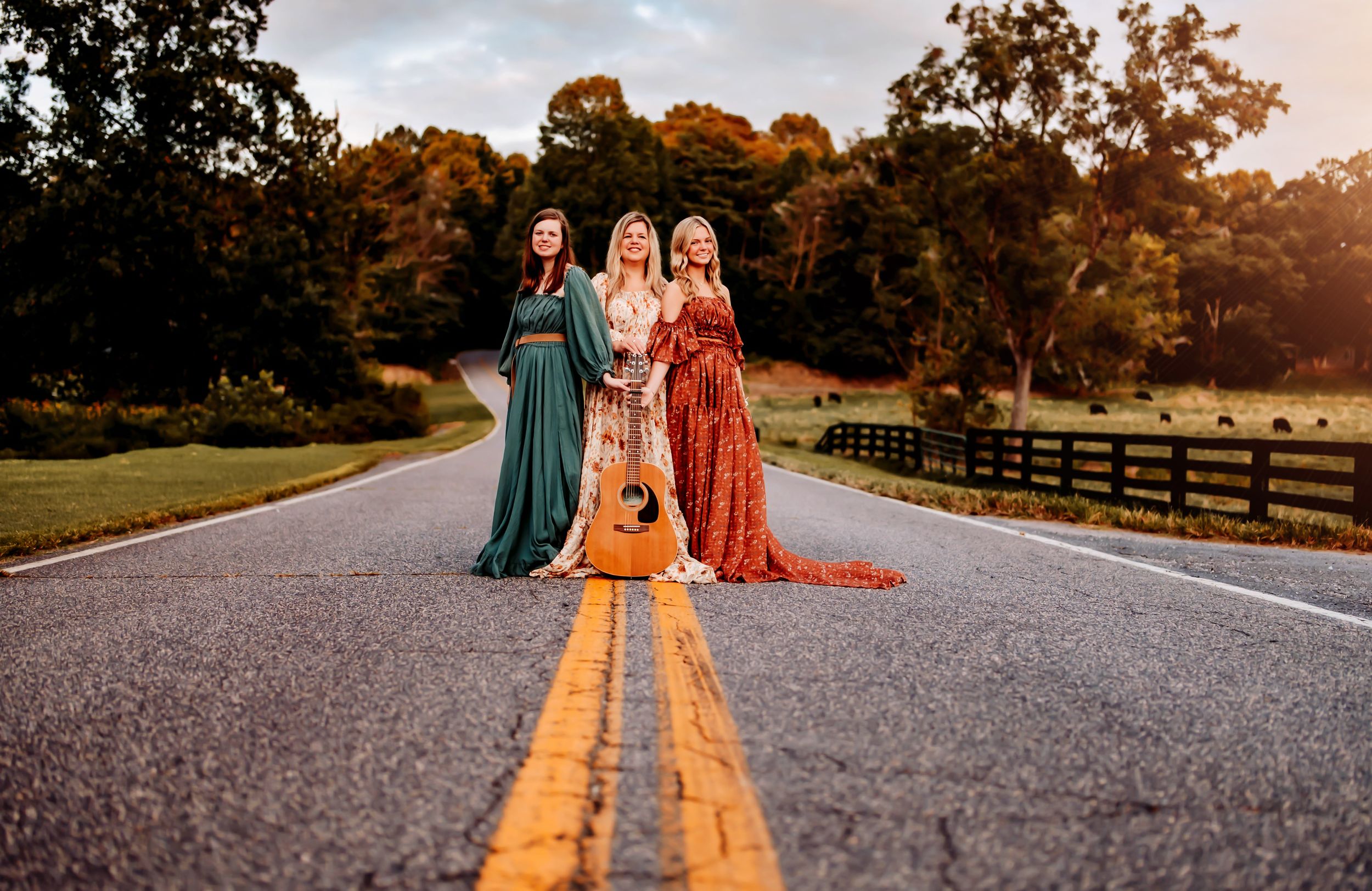 People in colorful dresses sit together in a golden sunset scene on a rustic gravel road amid fall foliage.