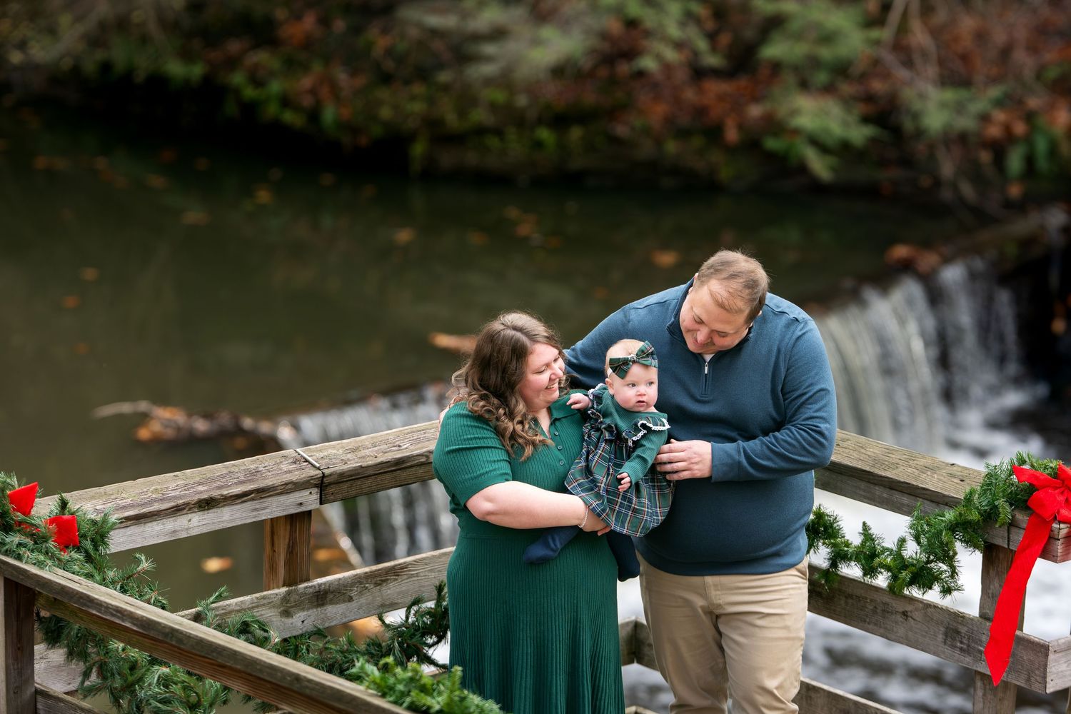 A couple admires their baby near a festive garland-decorated bridge with a waterfall backdrop.