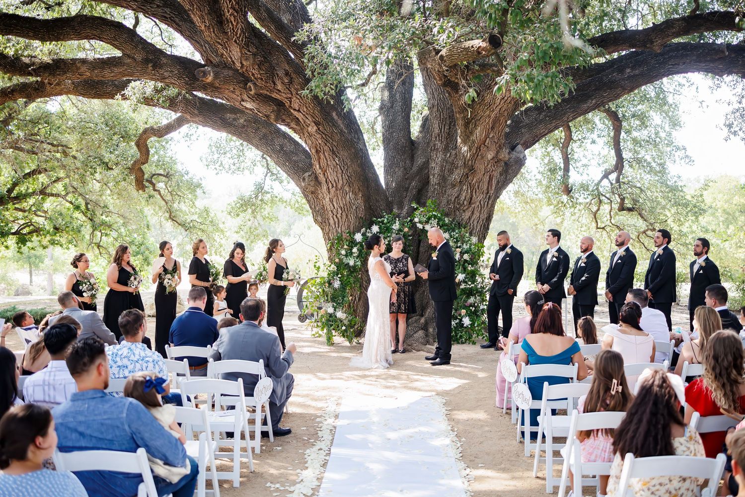 A wedding party in navy blue suits and white dresses walks together outside a church with arched doorways.