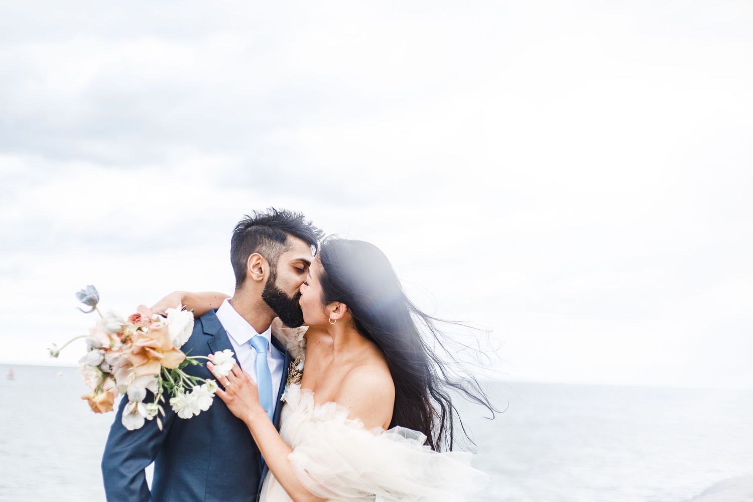 A couple in wedding attire embrace while holding a peach floral bouquet against a cloudy sky background.