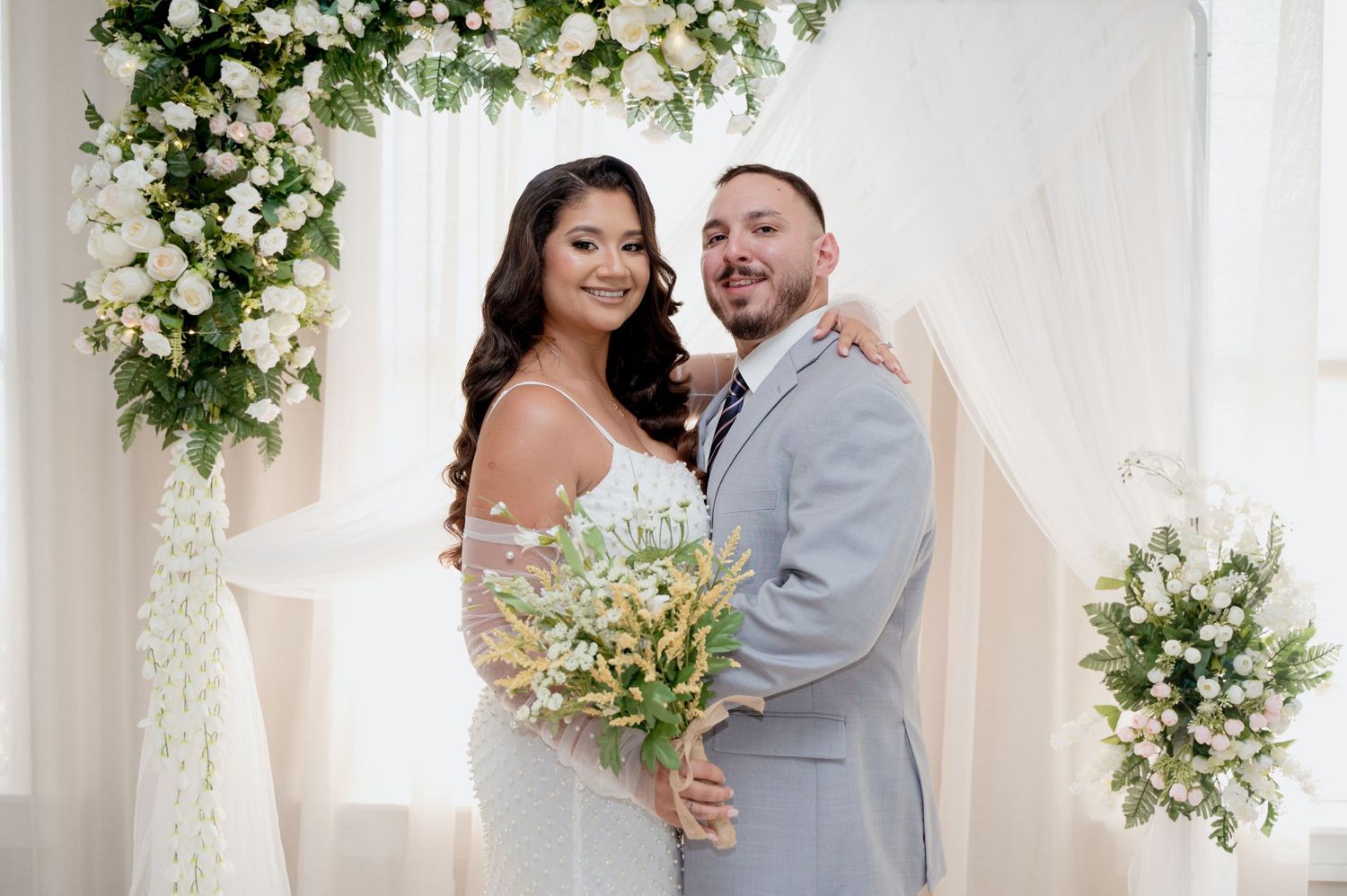 Couple shares romantic wedding moments under white floral arrangements in a bright indoor setting.