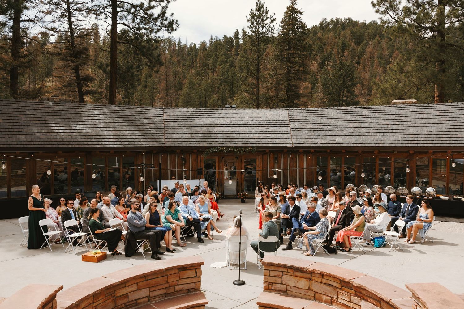 Outdoor wedding ceremony with guests seated in chairs at a rustic lodge surrounded by pine trees.