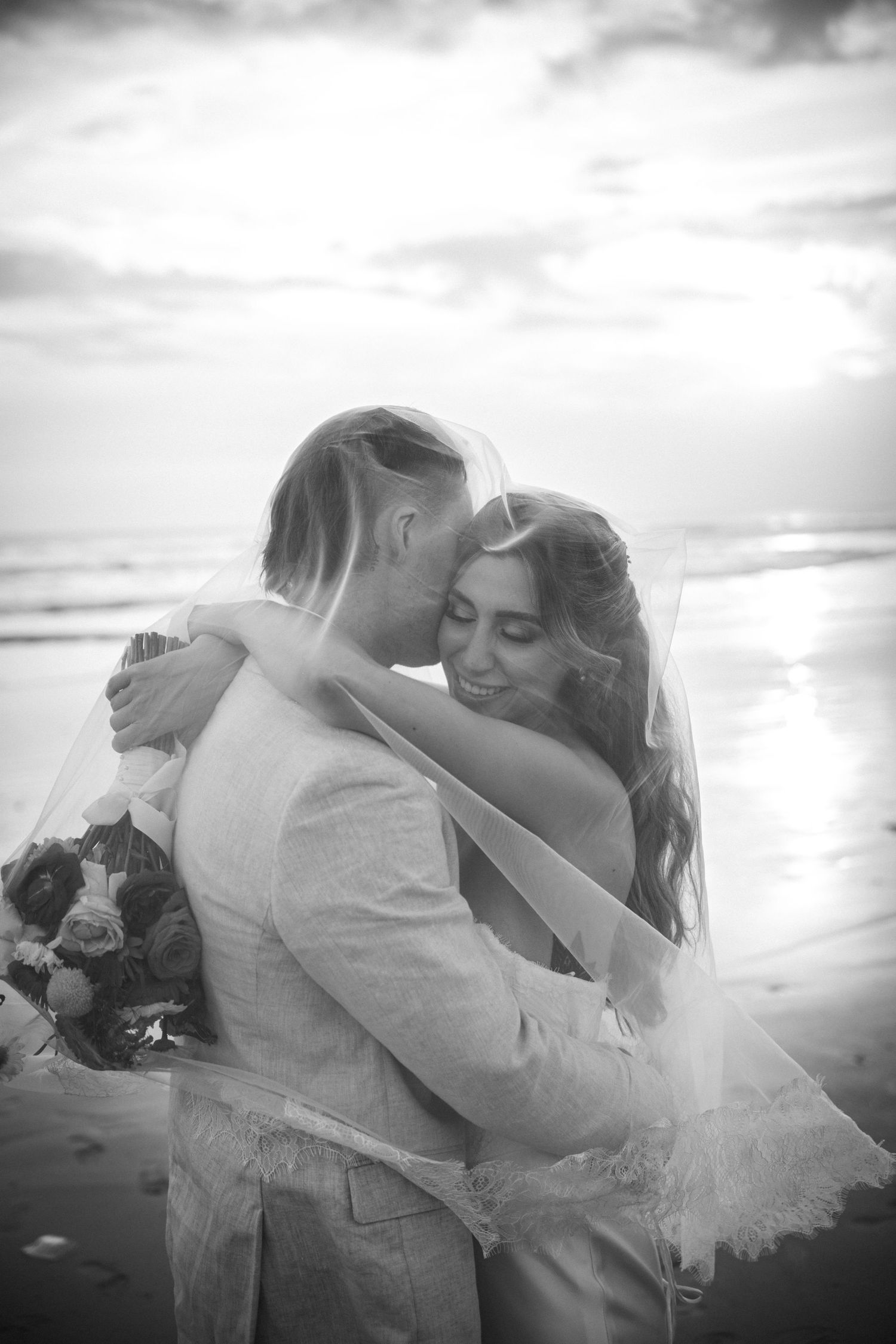 Silhouetted couple sharing a romantic kiss under flowing wedding veil at sunset with dramatic sky in background.