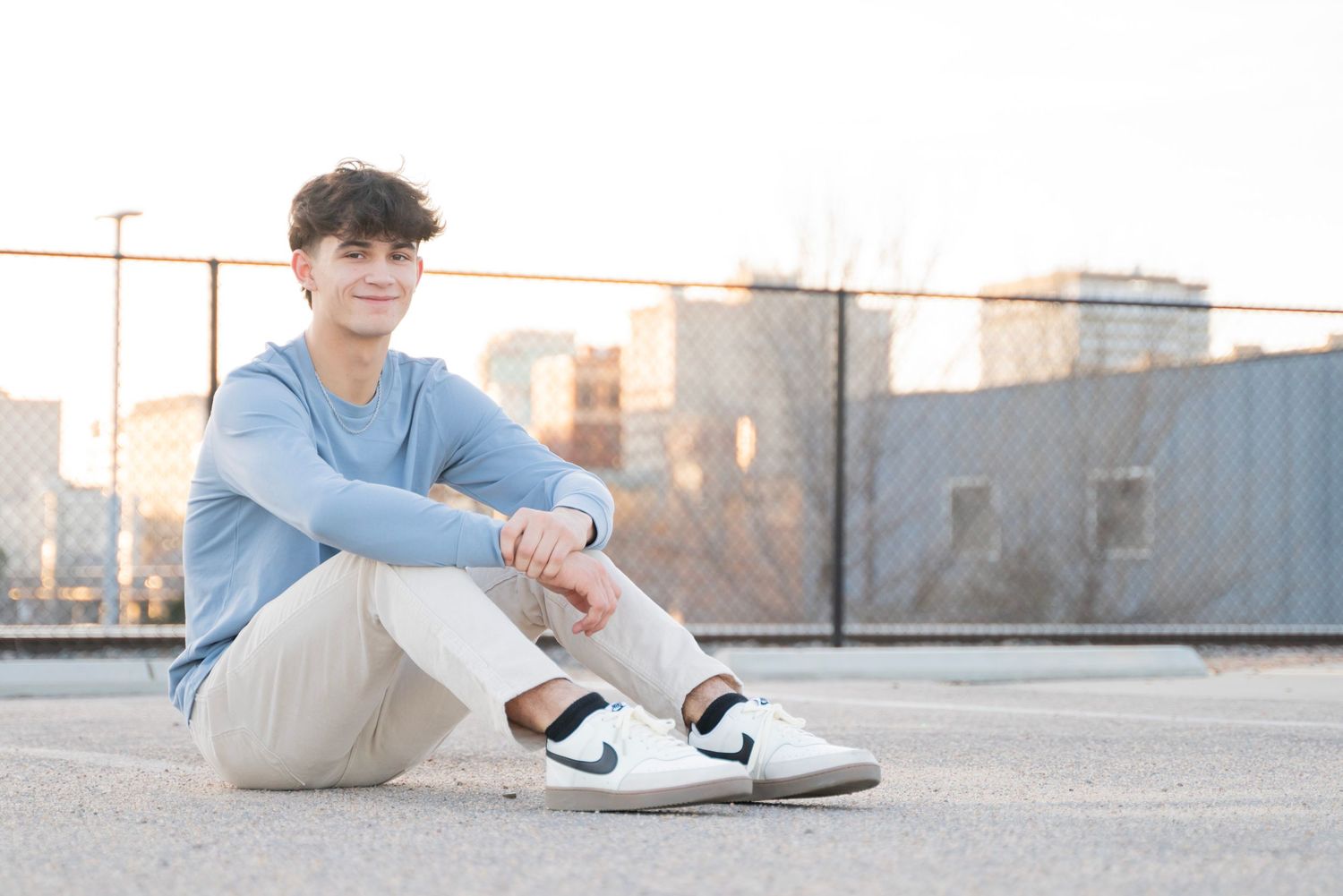 Smiling teenage boy in blue shirt and khakis sitting cross-legged on rooftop at sunset.